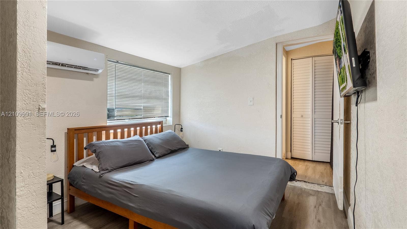 Room featuring wood-finish flooring, a wall-mounted climate control unit, a window with horizontal blinds, and a closet with louvered double doors