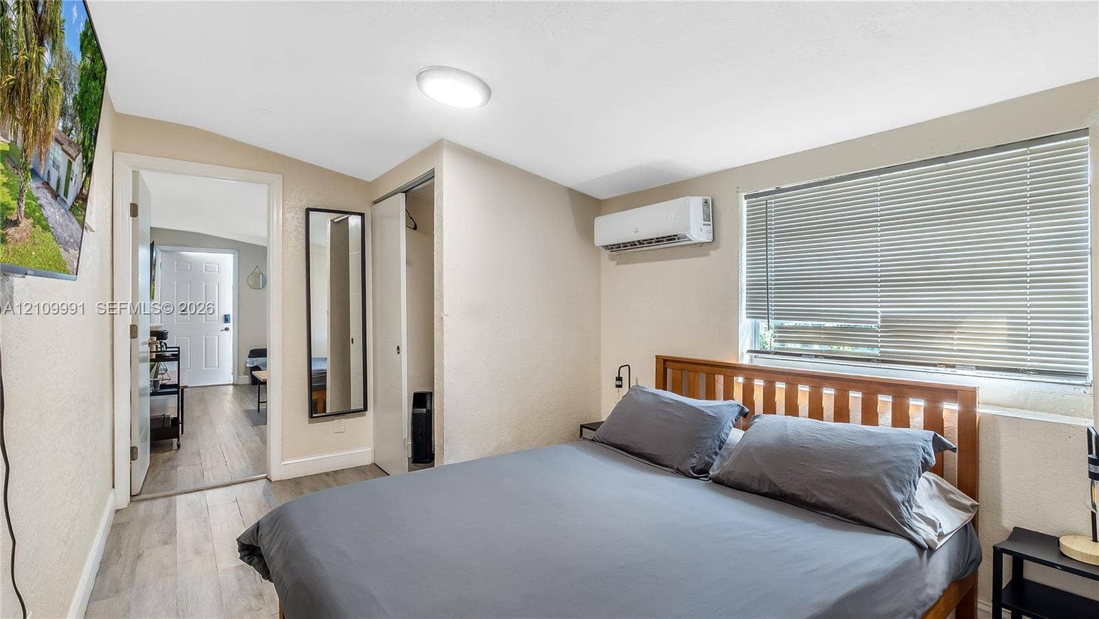 Neutral-toned room featuring wood-finish flooring, a wall-mounted ductless air conditioning unit, and a window with horizontal blinds