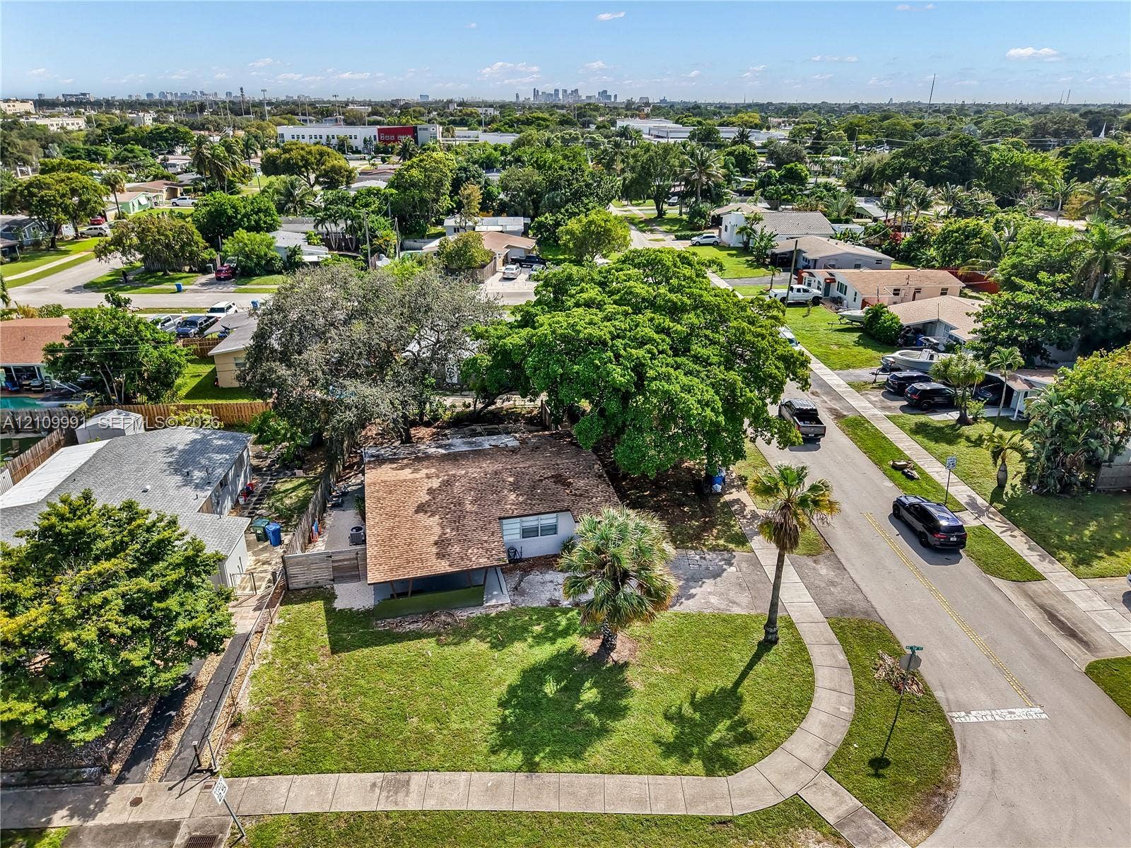 Aerial perspective featuring a single-story residence with a shingled roof, corner lot placement, mature shade trees, and manicured lawn