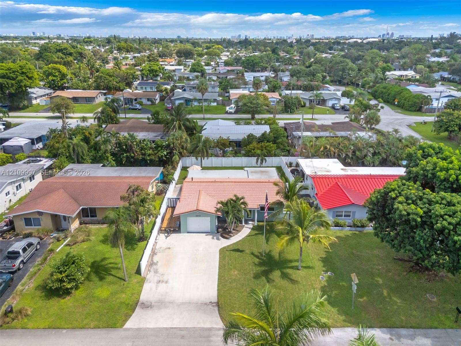 Single-story residence featuring a concrete tile roof, attached one-car garage, concrete driveway, white privacy fencing, and mature palm trees