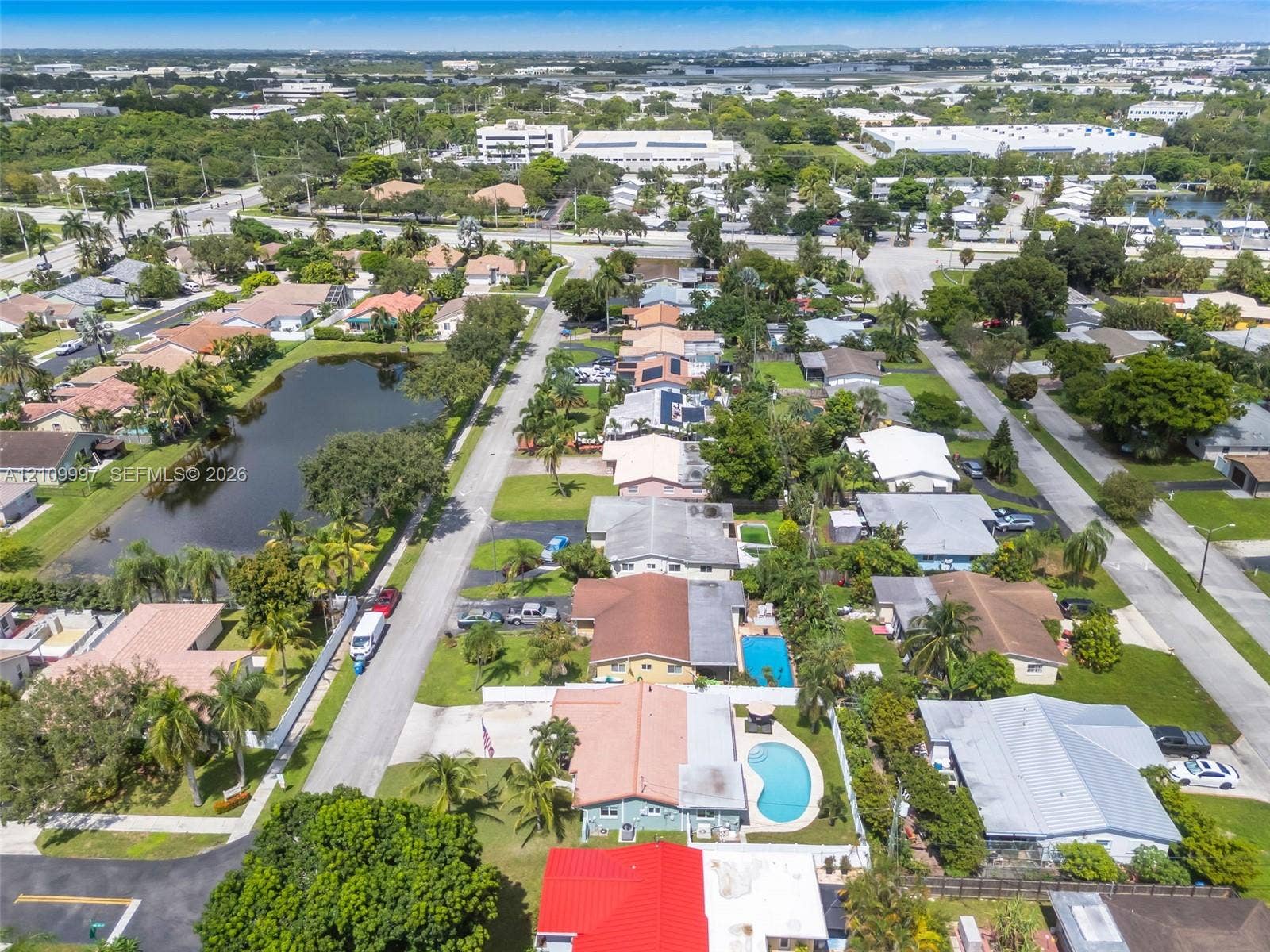 Aerial perspective of a residential neighborhood featuring paved roadways, established landscaping with mature palm trees, single-story residences with diverse rooflines, and localized water features