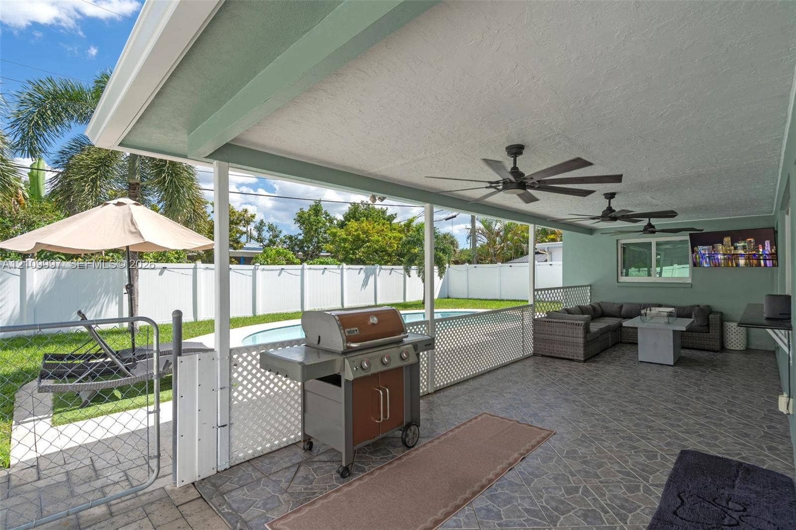 Covered patio featuring stone-patterned tile flooring, recessed ceiling fans, and a white lattice privacy screen, opening to an outdoor area with a swimming pool and vinyl perimeter fencing