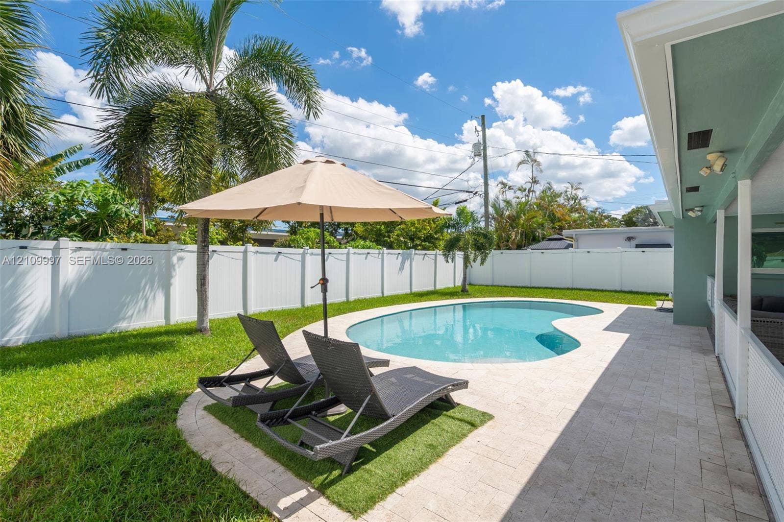 Private backyard featuring a freeform swimming pool, expansive stone-paver patio, white vinyl perimeter fencing, and mature palm trees