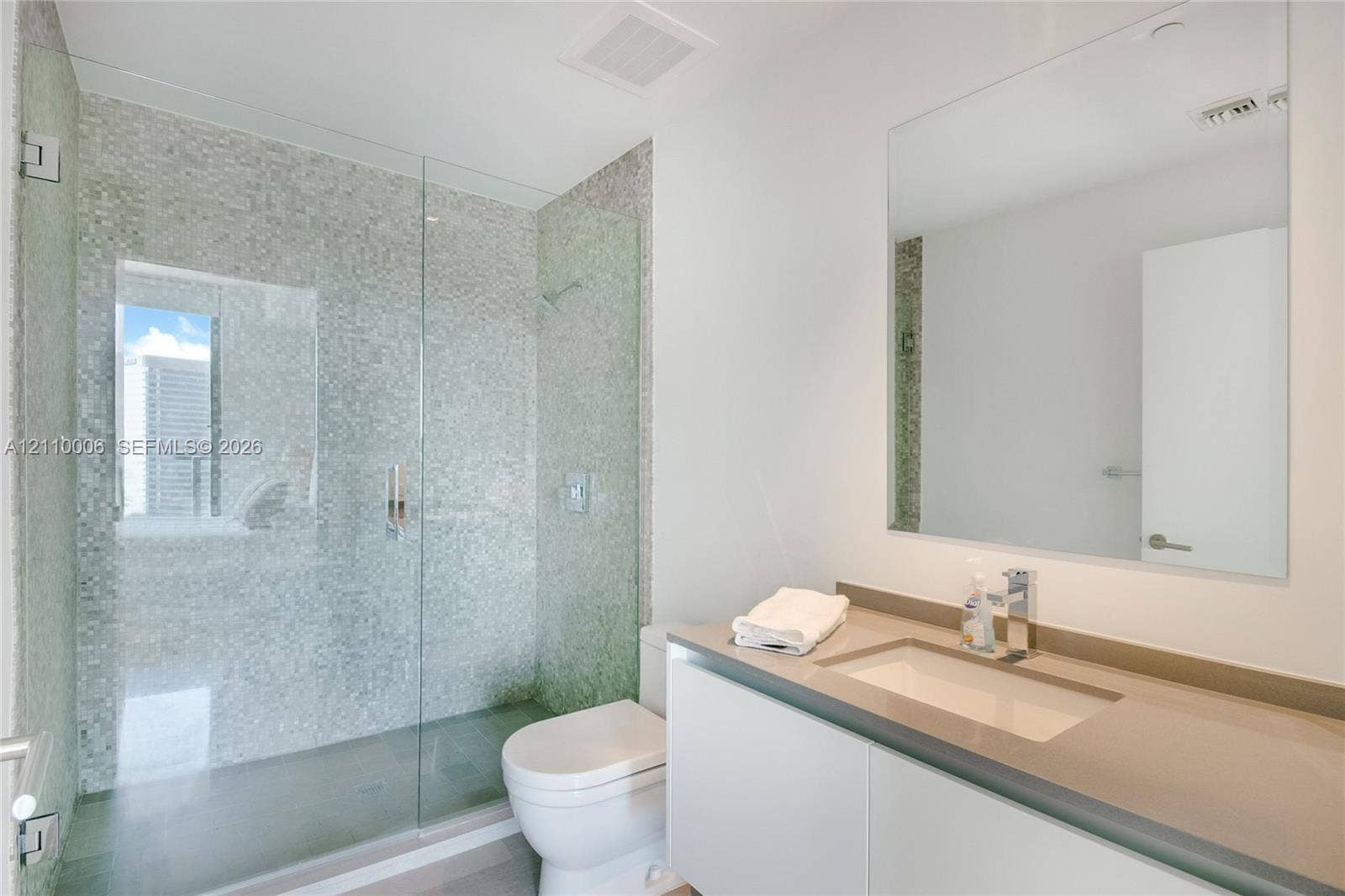 Walk-in shower featuring a glass enclosure and metallic mosaic tile surround, white wall-mounted vanity, rectangular undermount sink, solid surface countertop, and large wall-mounted mirror