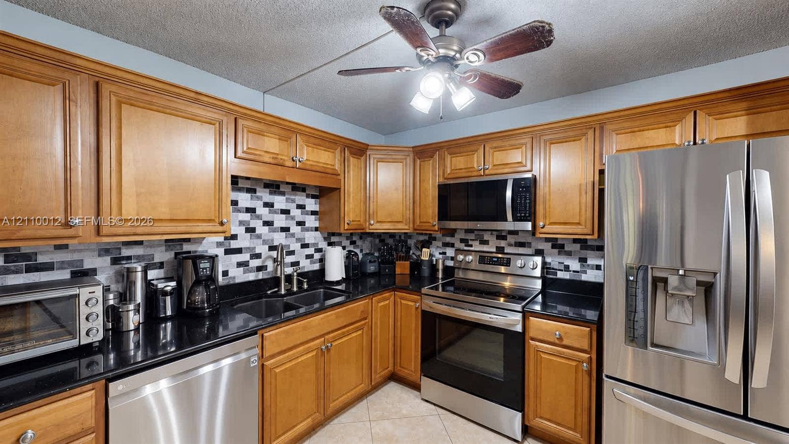 1601 16th Terrace, Palm Beach Gardens, FL 33418 Kitchen featuring natural finish wood cabinetry, contrasting black countertops, a multi-tonal rectangular tile backsplash, stainless steel appliances, and a ceiling-mounted fan with integrated lighting