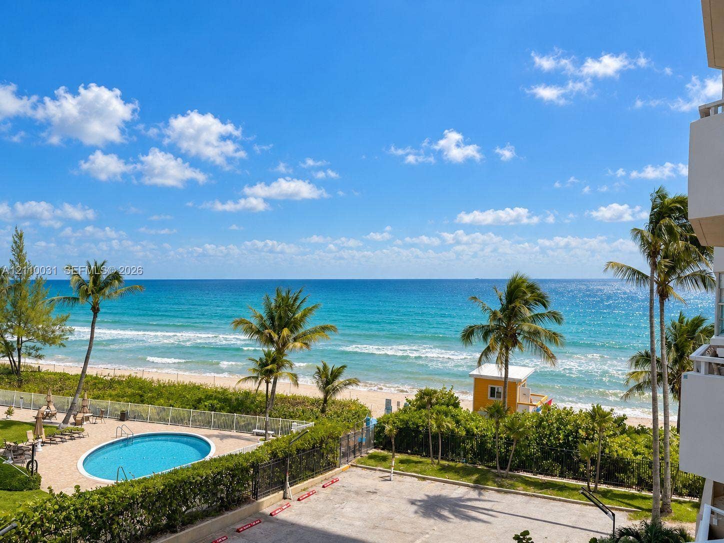 Oceanfront perspective featuring a swimming pool, paved surface parking, perimeter fencing, and mature palm trees