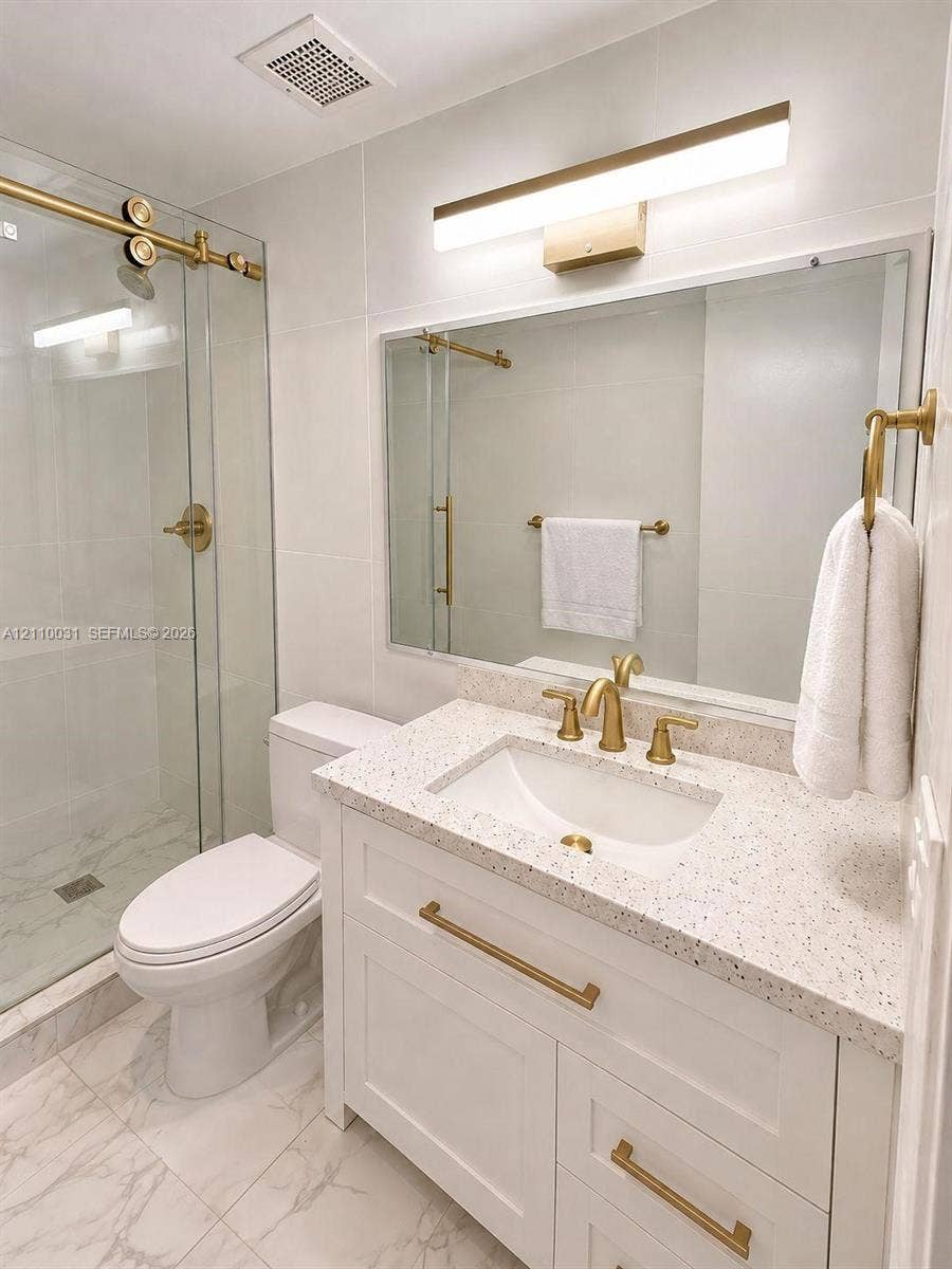 Bathroom featuring a white vanity with speckled countertop, gold-toned hardware, glass-enclosed shower, and large-format tile flooring