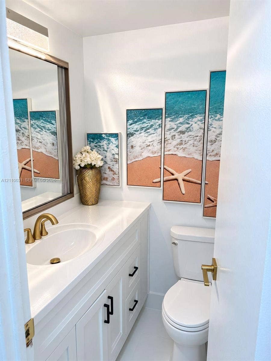 Vanity with white solid-surface countertop and undermount sink, brass-finish widespread faucet, white shaker-style cabinetry with matte black hardware, and rectangular vanity mirror with integrated light fixture