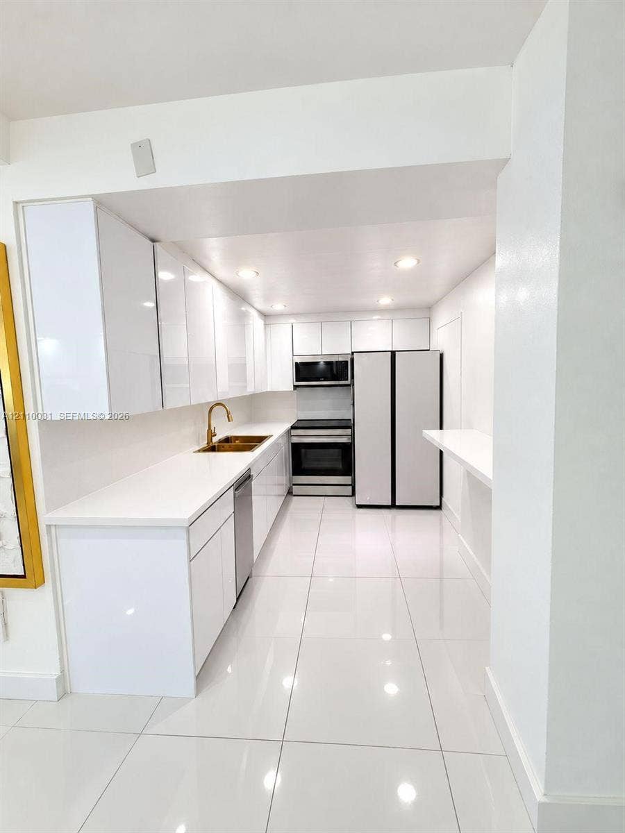 Kitchen featuring high-gloss white cabinetry, quartz countertops, metallic-finish faucet, stainless steel appliances, and large-format tile flooring