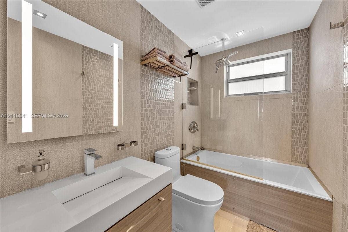 1610 Lenox Avenue, #516, Miami Beach, FL 33139 Bathroom featuring a wood-finish vanity with a white rectangular vessel sink, a wall-mounted LED-backlit mirror, a glass-enclosed shower and tub combination, and neutral textured tile wall accents