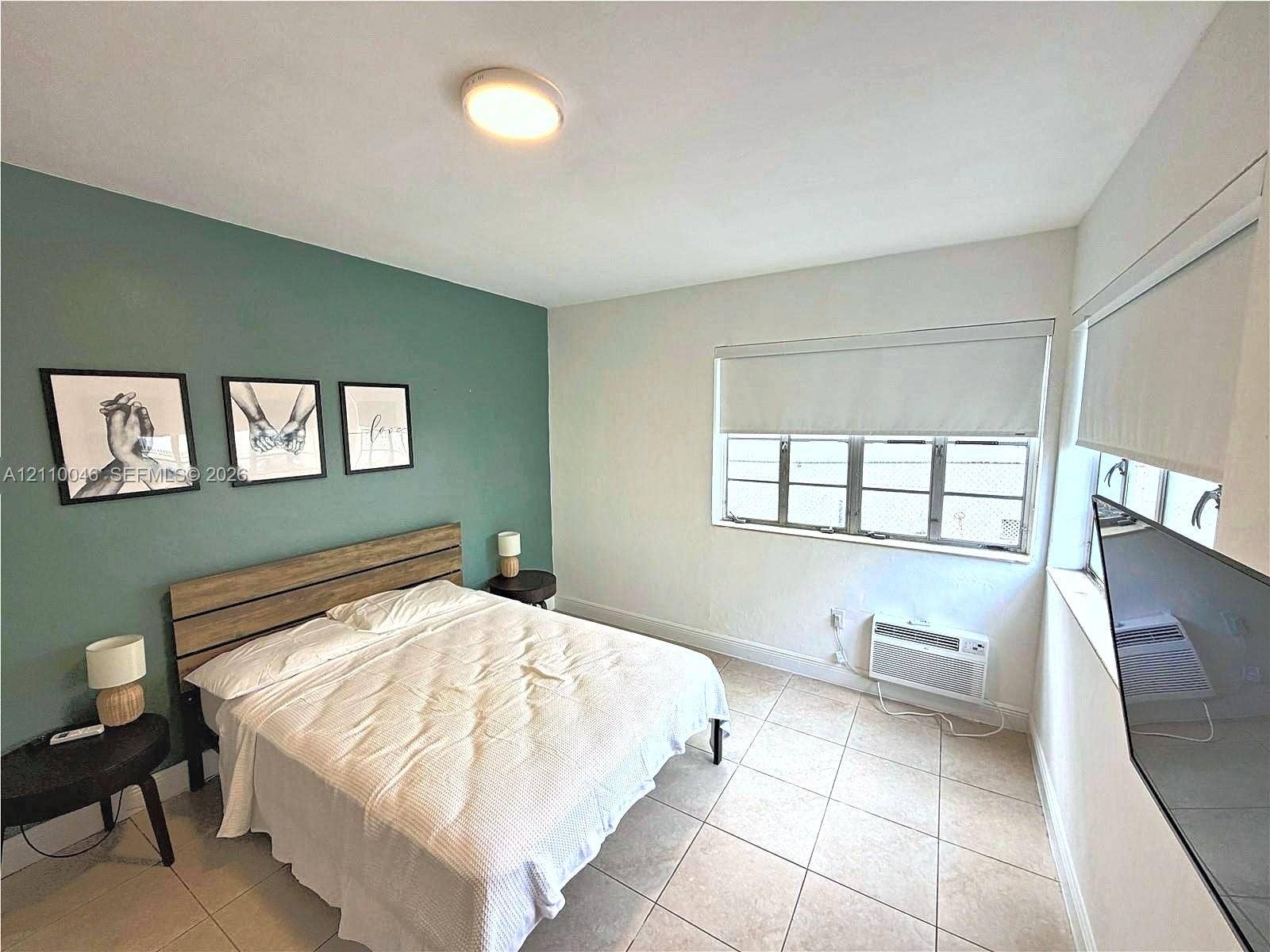 Neutral-tone tiled flooring, accent wall in muted sage, multi-pane window with installed roller shade, and wall-mounted air conditioning unit