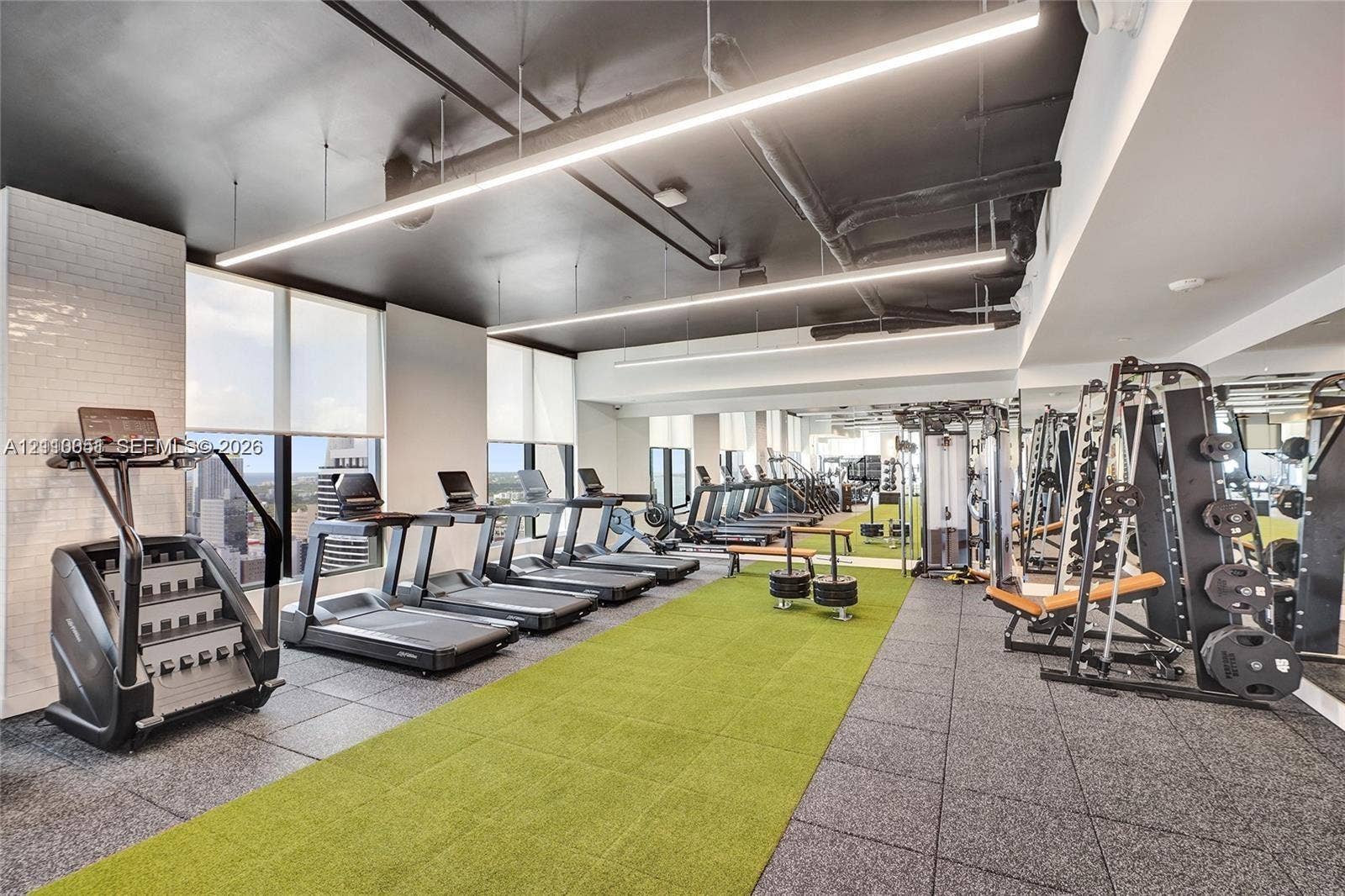 121 NE 5th Street, #1703, Miami, FL 33132 Fitness facility featuring floor-to-ceiling windows, high-contrast exposed industrial-style ceiling, integrated linear LED lighting, wall-to-wall mirrored surfaces, and a combination of rubber and artificial turf flooring