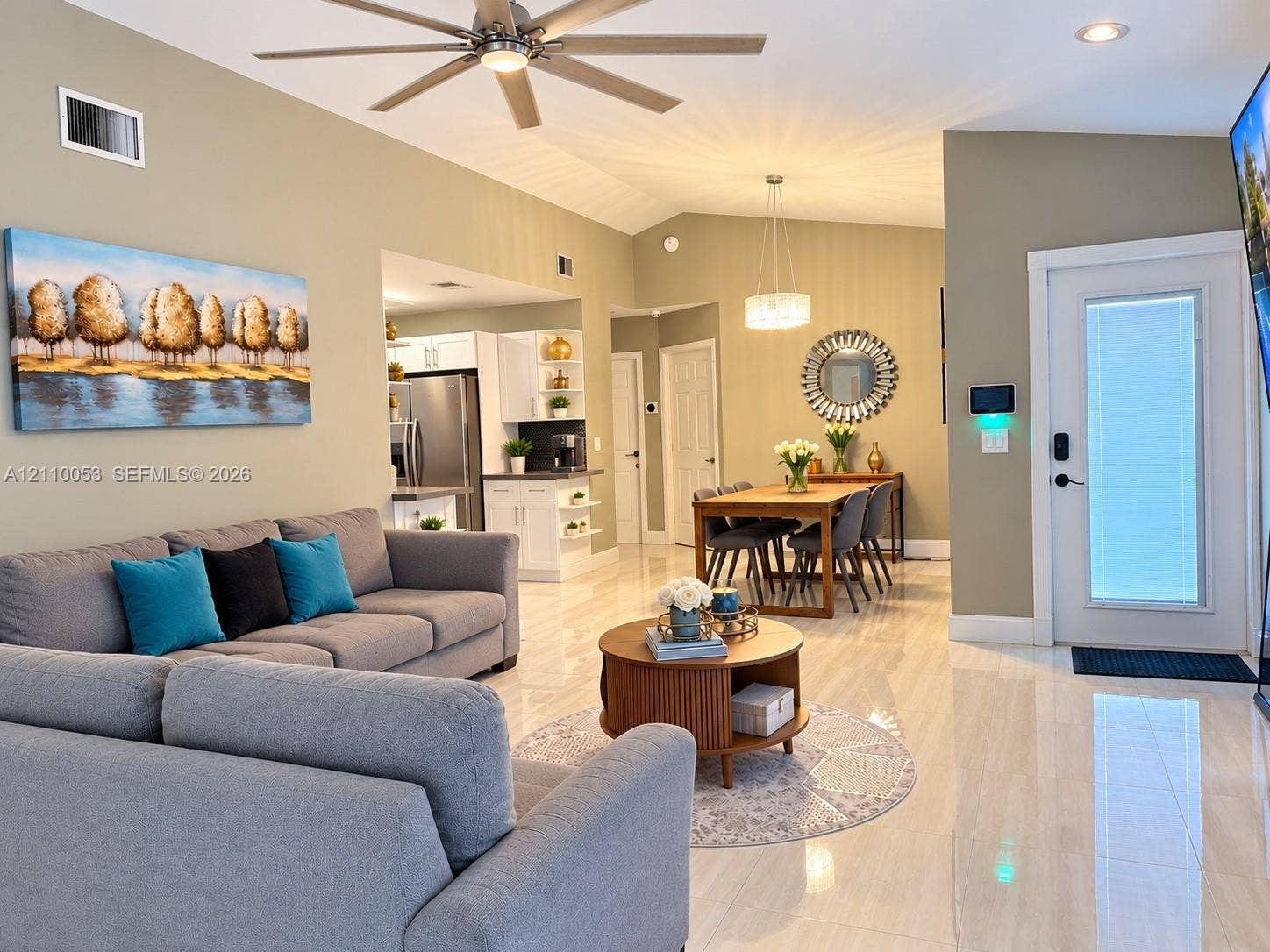 Open-concept interior featuring large-format tile flooring, vaulted ceilings, a ceiling fan with integrated lighting, and a modern exterior door with an inset glass panel