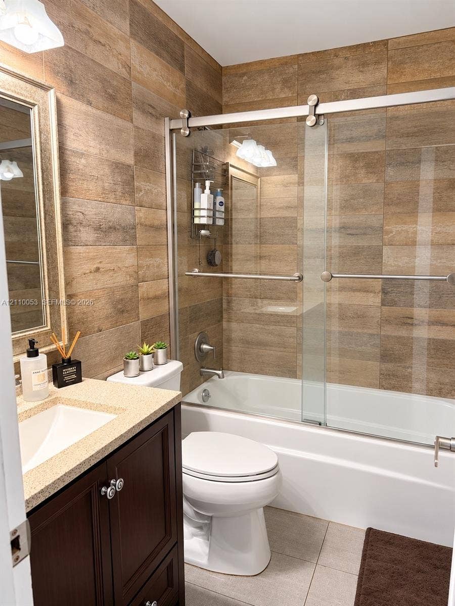 Bathroom featuring wood-finish wall tile, a dark wood-finish vanity with a stone-composite countertop, and a glass-enclosed shower and tub combination