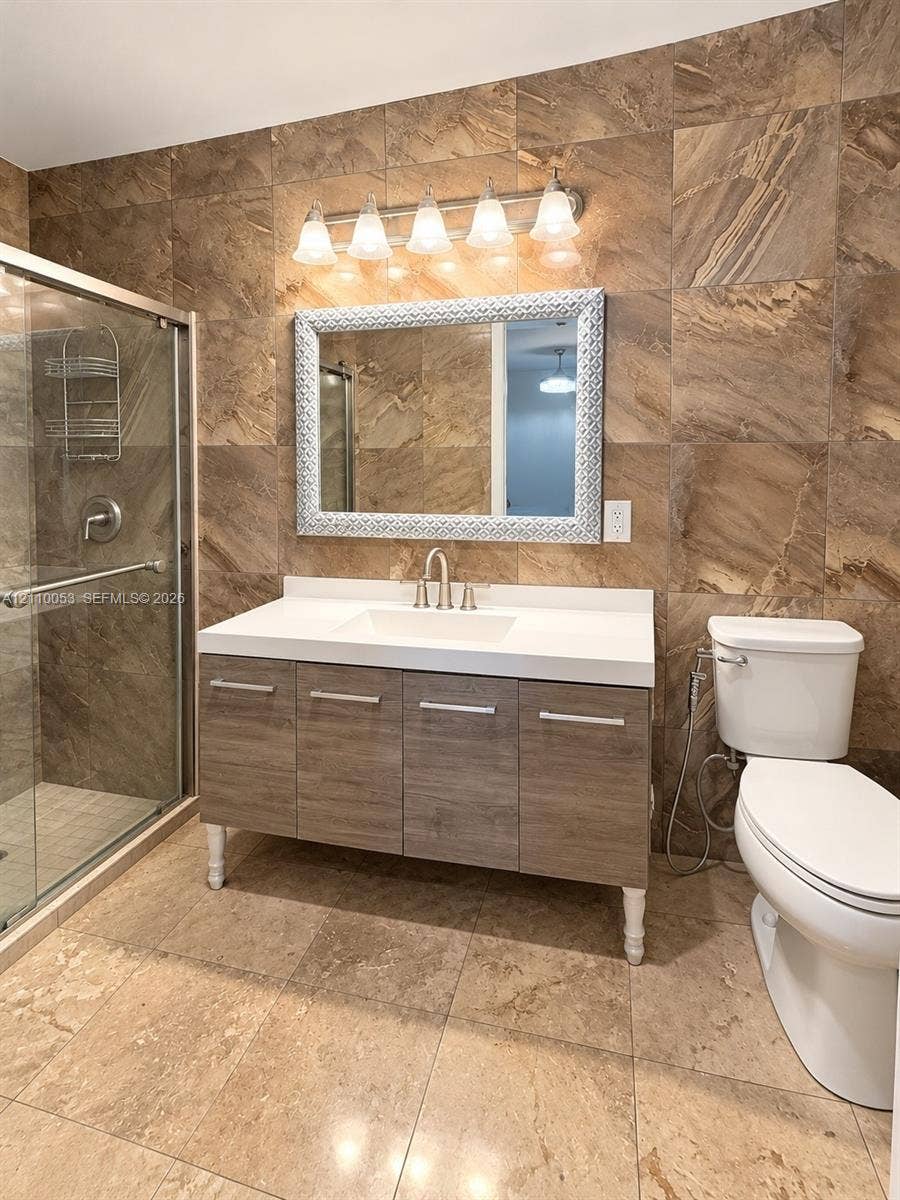 Bathroom featuring a vanity with a white composite countertop, wood-finish cabinetry, floor-to-ceiling large-format tile walls, tile flooring, and a glass-enclosed shower
