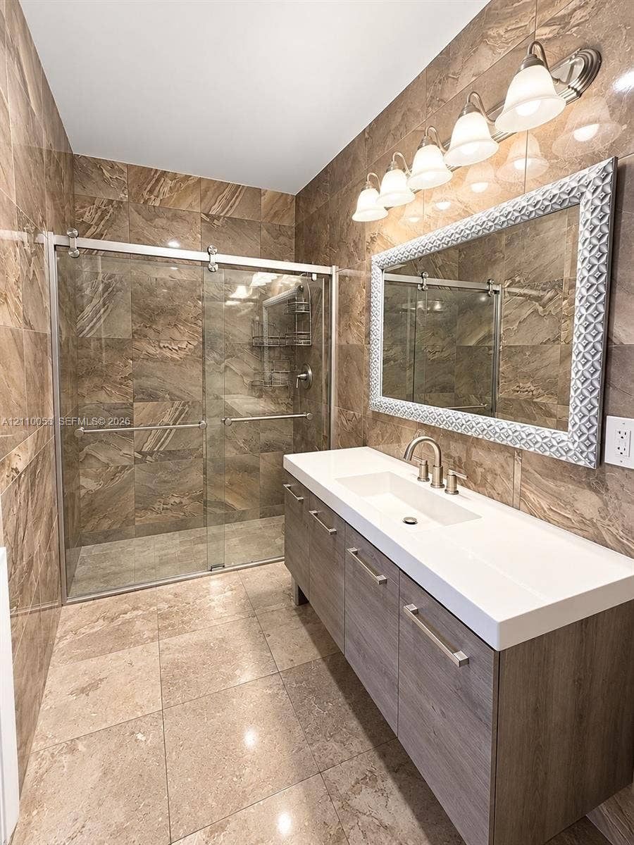 Bathroom featuring floor-to-ceiling stone-tile walls, a glass-enclosed shower with sliding doors, a vanity with a white integrated basin, and wood-finish cabinetry
