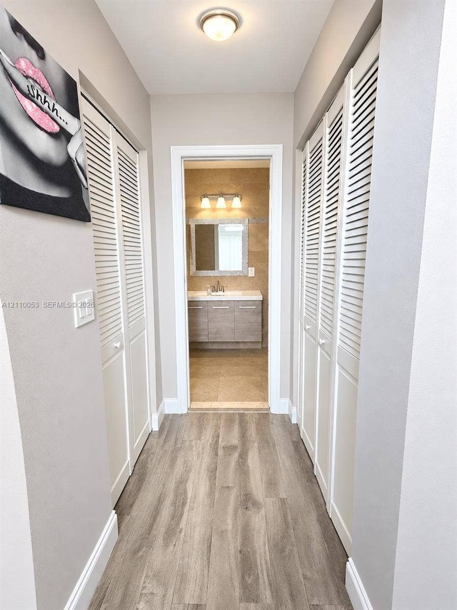 Hallway featuring dual sets of white louvered closet doors, wood-finish flooring, and a vanity area equipped with a mirror, three-light fixture, and stone-finish cabinetry