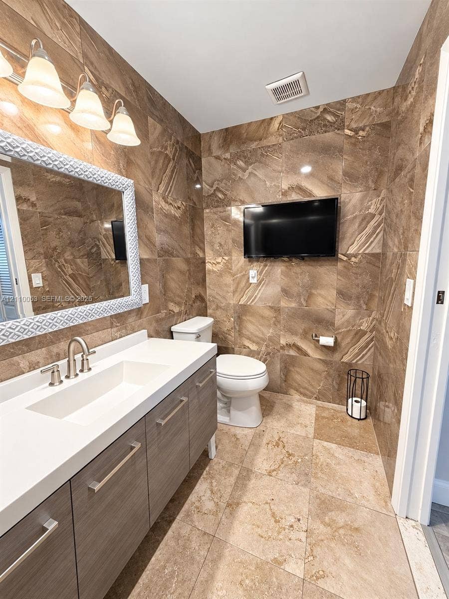 Bathroom featuring full-height large-format stone tile walls, floor-to-ceiling stone tile flooring, a flat-panel wood-finish vanity with a white integrated countertop and sink, and a multi-bulb vanity light fixture
