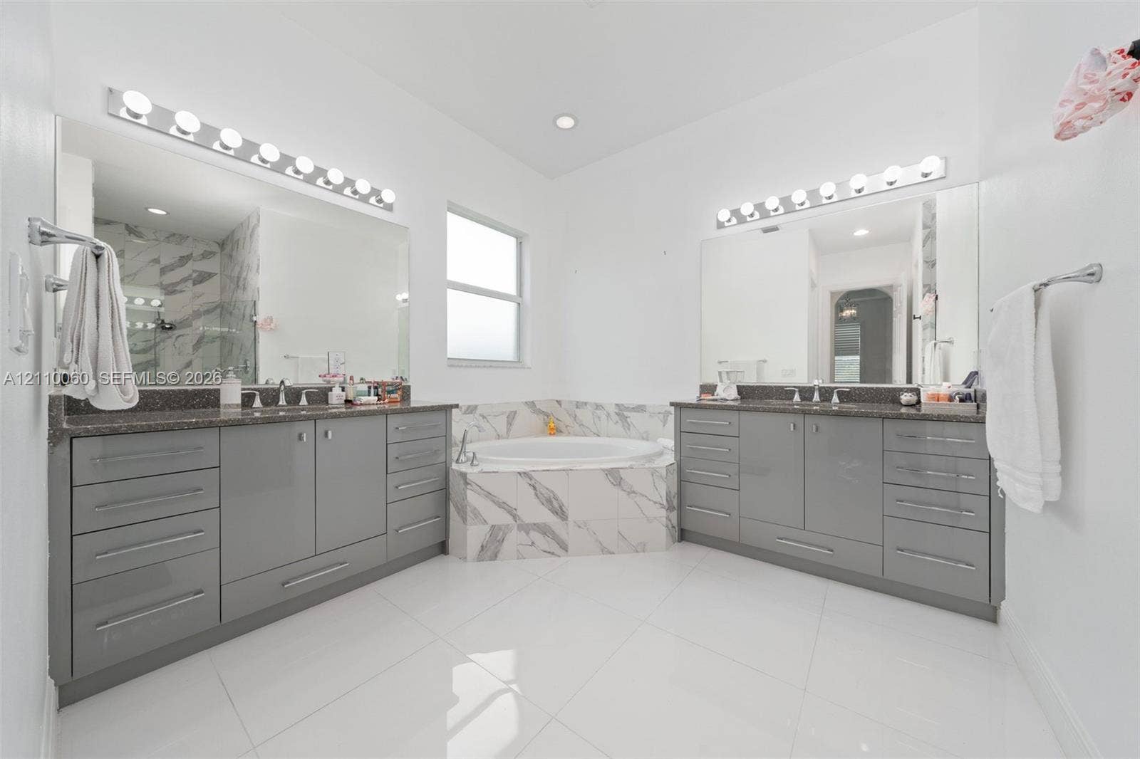 Dual vanities featuring gloss-finish cabinetry, stone countertops, integrated basins, and polished hardware, complemented by a tiled soaking tub and porcelain tile flooring