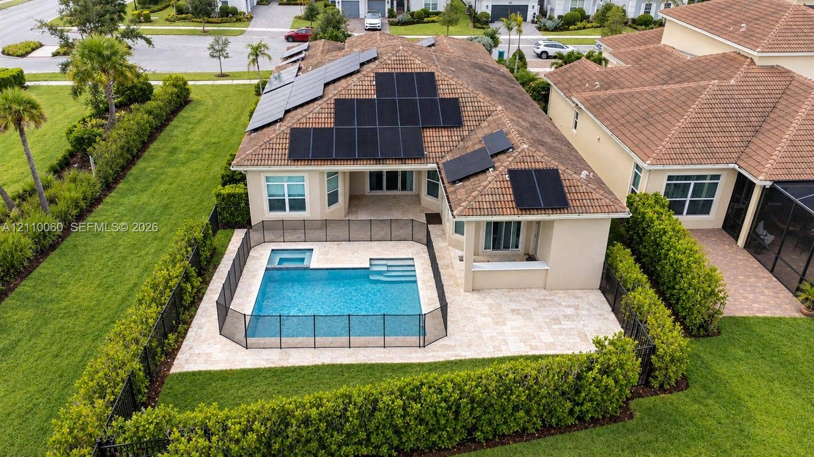 Aerial view of a residential property featuring a tiled roof equipped with integrated solar panels, a private swimming pool with a built-in spa, and a stone-paver patio enclosed by a perimeter fence