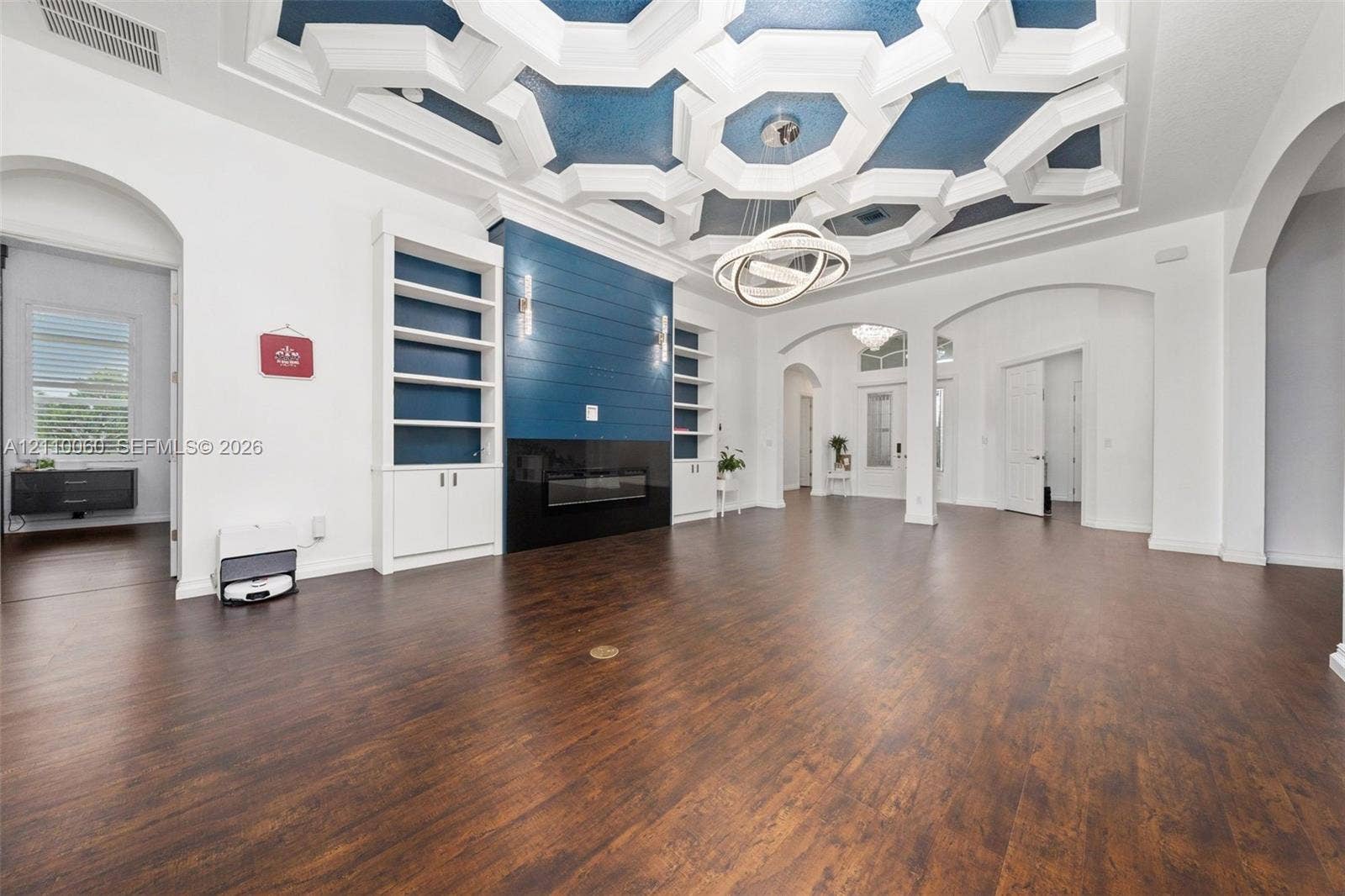 Geometric coffered ceiling with inset blue accents, wood-finish flooring, built-in floor-to-ceiling cabinetry, shiplap fireplace surround, and modern circular tiered chandelier