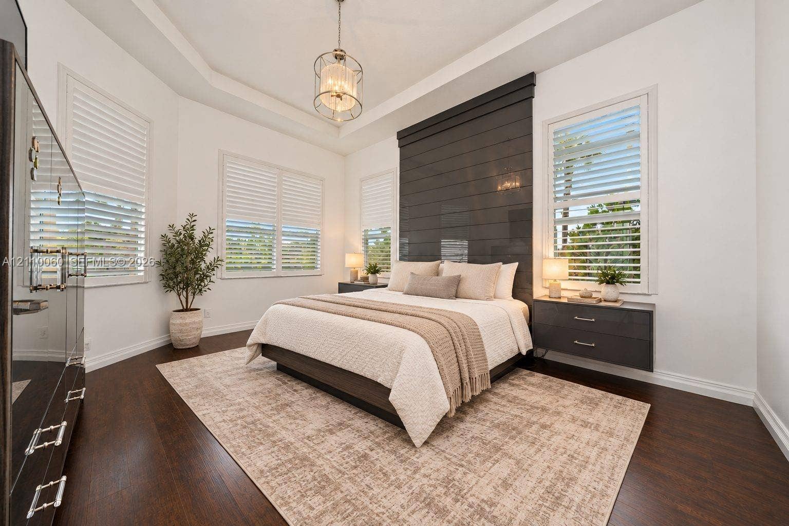 Virtually staged. Tray ceiling with a central pendant light, wood-finish flooring, charcoal horizontal plank accent wall, and dual floating bedside drawers