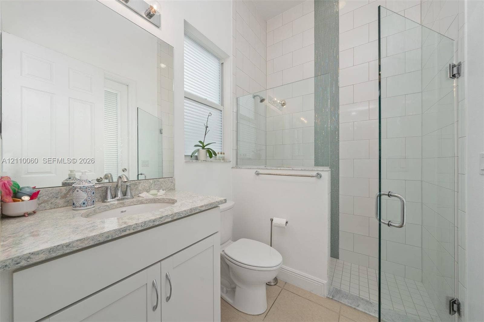 Bathroom featuring a vanity with stone-finish countertop and undermount sink, large wall-mounted mirror, walk-in shower with glass enclosure, vertical tile accent, and light-tone tile flooring