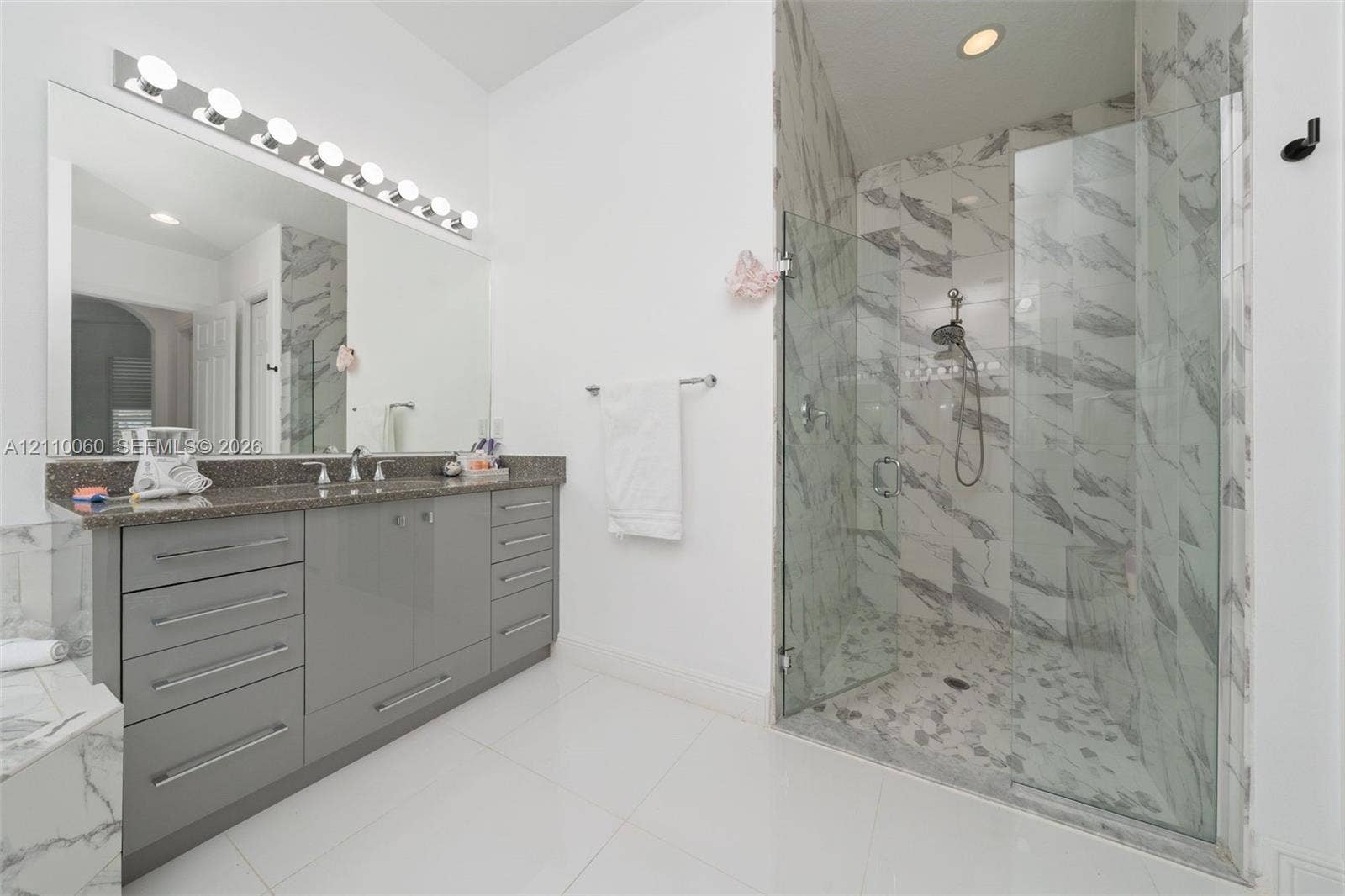 Bathroom featuring a dual-sink vanity with grey cabinetry, a solid-surface countertop, full-width mirror with multi-bulb lighting, and a glass-enclosed shower with tiled wall surrounds