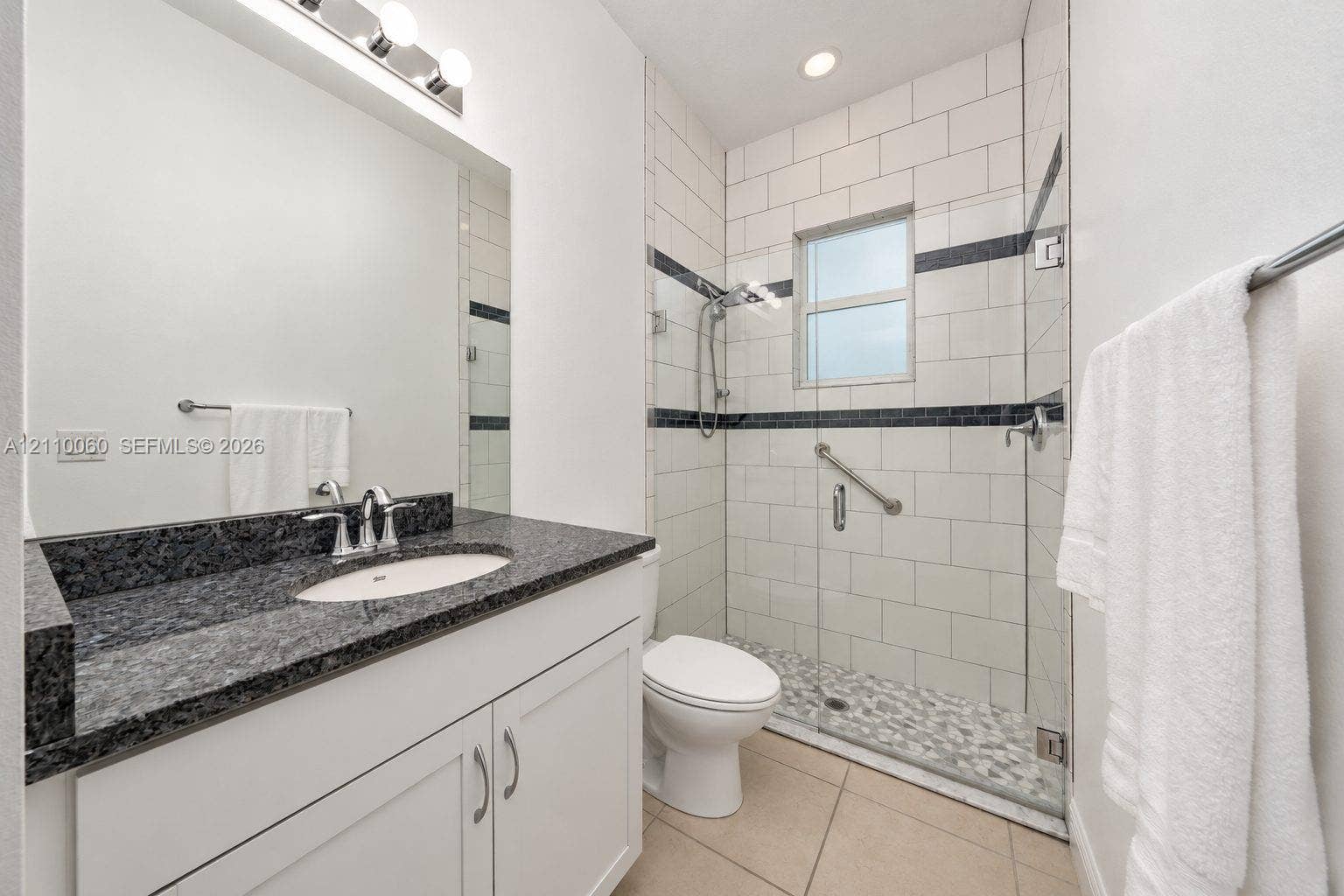 Bathroom featuring a white vanity cabinet with a dark stone countertop, an integrated sink with a chrome faucet, a walk-in shower with a glass enclosure and multi-tone tile accents, and large-format tile flooring
