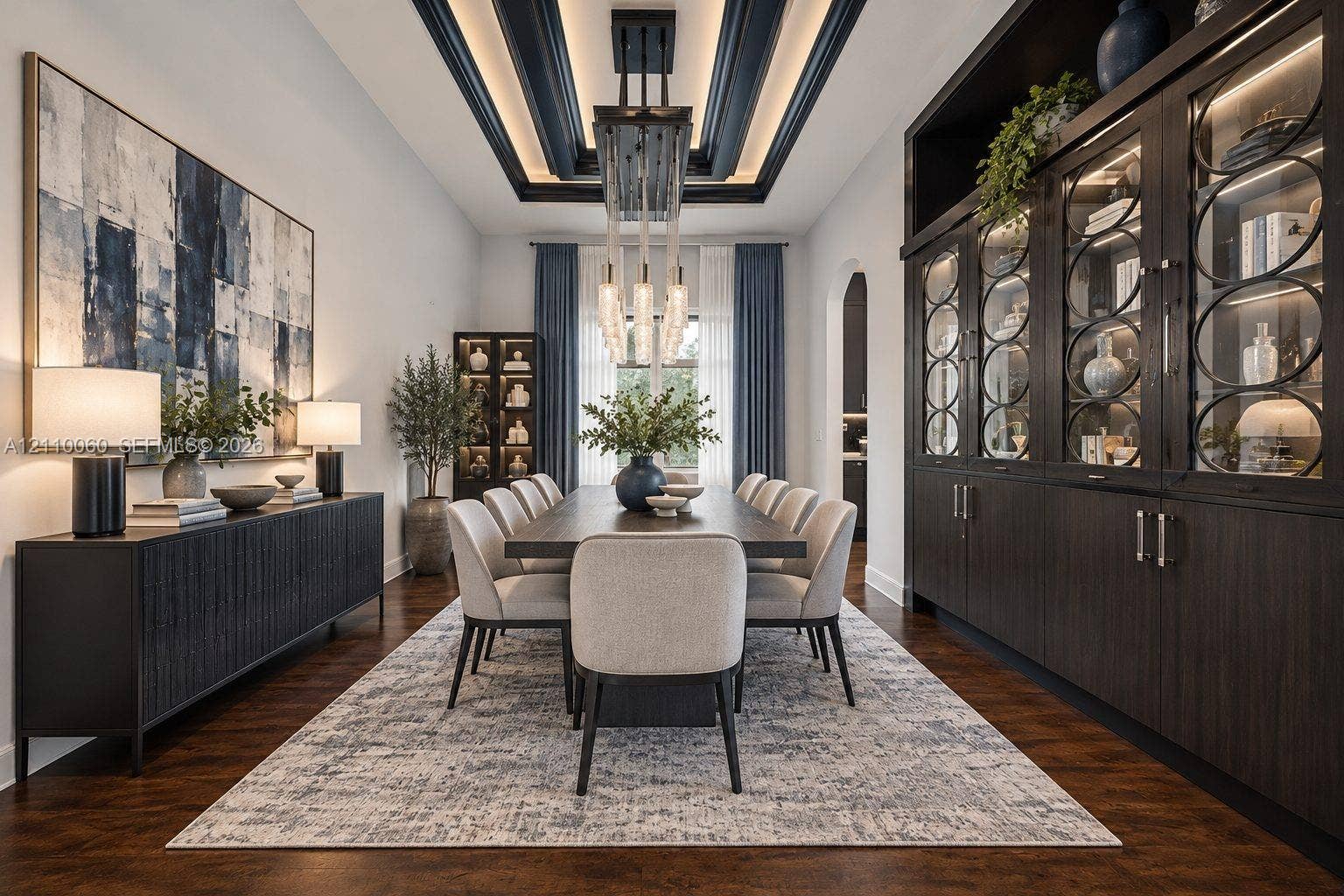 Virtually staged. Formal dining area featuring a tray ceiling with recessed lighting, custom floor-to-ceiling built-in cabinetry with glass-paneled display doors, dark wood-finish flooring, and a multi-tiered crystal pendant light fixture