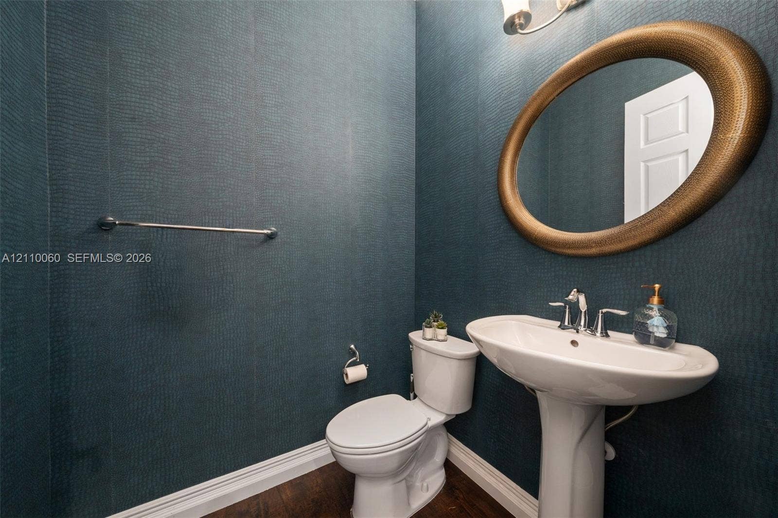 Textured deep-teal wall covering, white pedestal sink with chrome faucet, oval wall-mounted mirror with bronze finish, and dark wood-finish flooring
