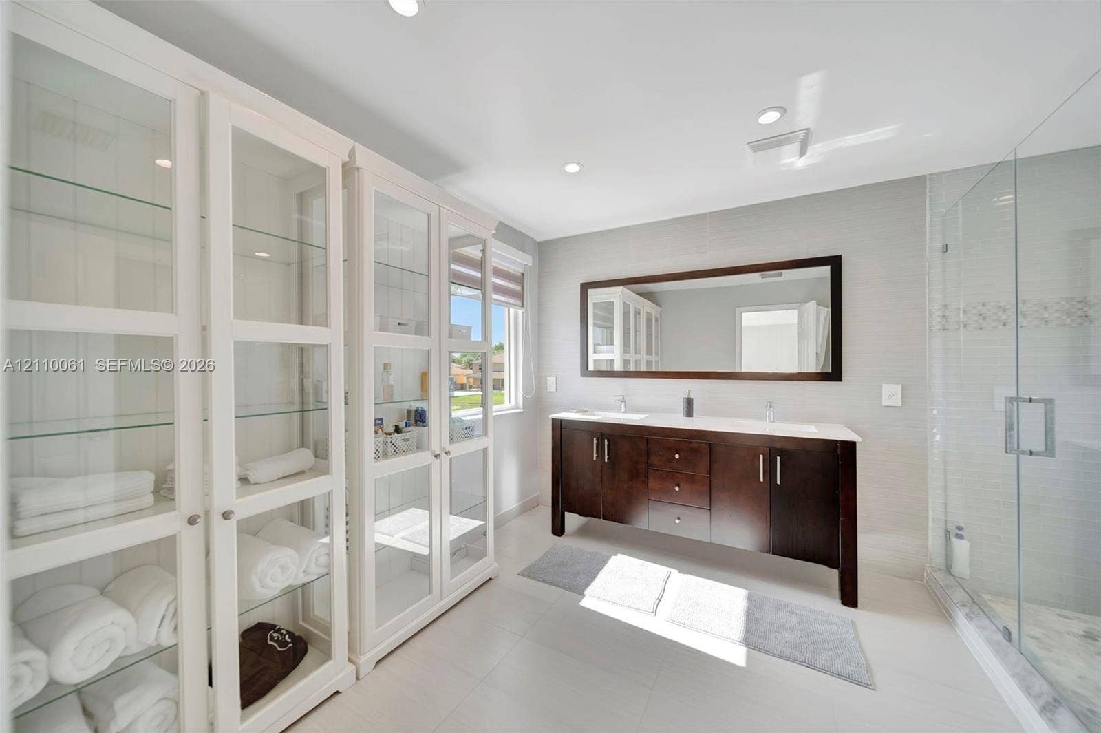 4424 E Whitewater Avenue, Weston, FL 33332 Double vanity with a dark wood-finish cabinet and surface countertop, glass-enclosed shower with tile surround, and extensive built-in cabinetry featuring glass-front doors