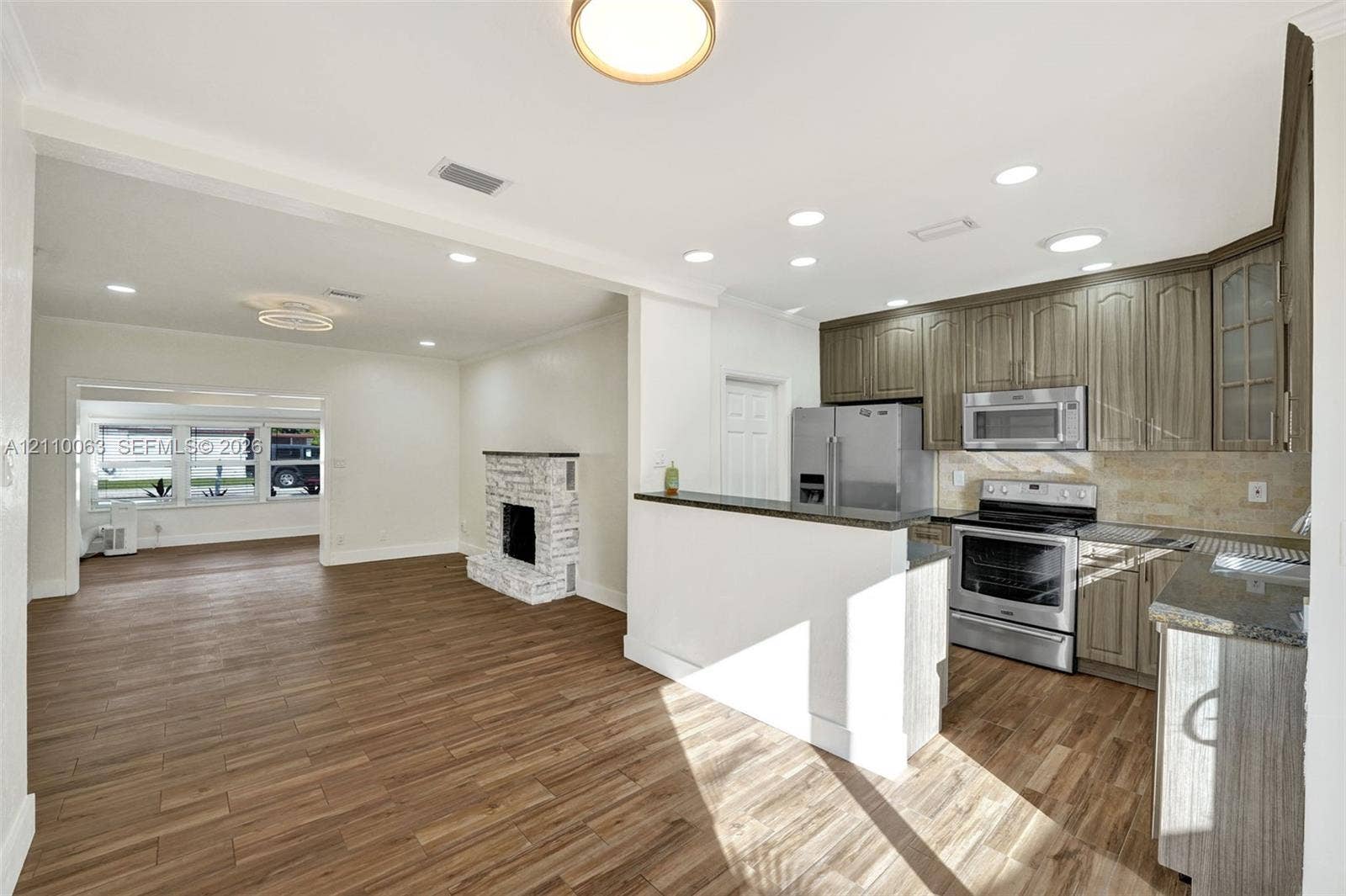 Kitchen featuring wood-finish cabinetry, stone-texture backsplash, stainless steel appliances, and a breakfast bar, complemented by wood-finish flooring and a whitewashed brick fireplace