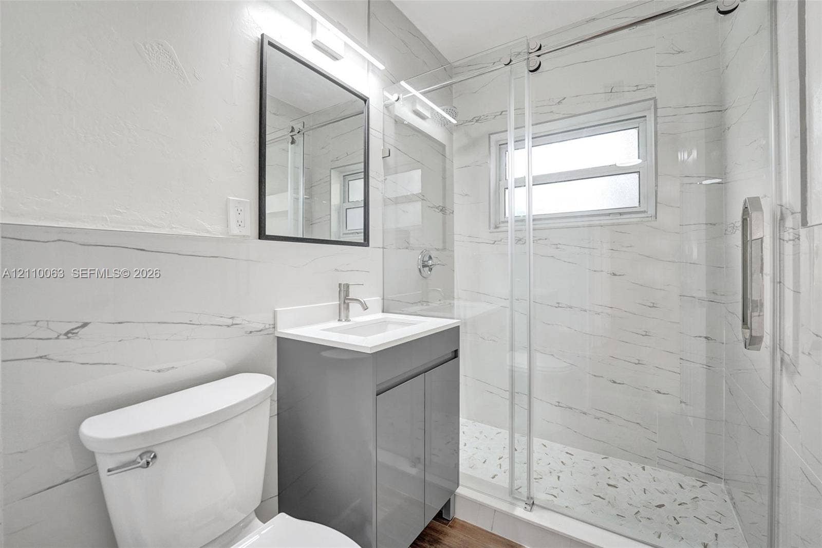 Bathroom featuring full-height large-format wall tile, glass-enclosed shower, stone-finish vanity with rectangular basin, and black-framed wall mirror