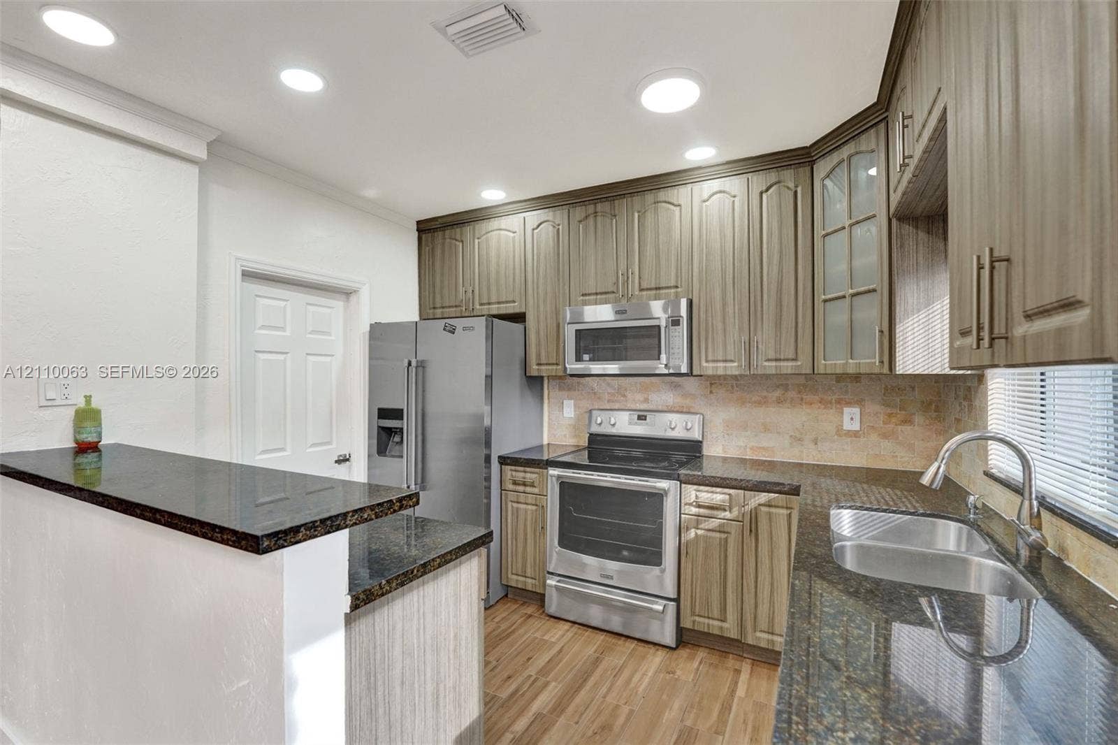 Kitchen featuring wood-finish cabinetry, dark stone countertops, stainless steel appliances, a tiled backsplash, and wood-finish flooring