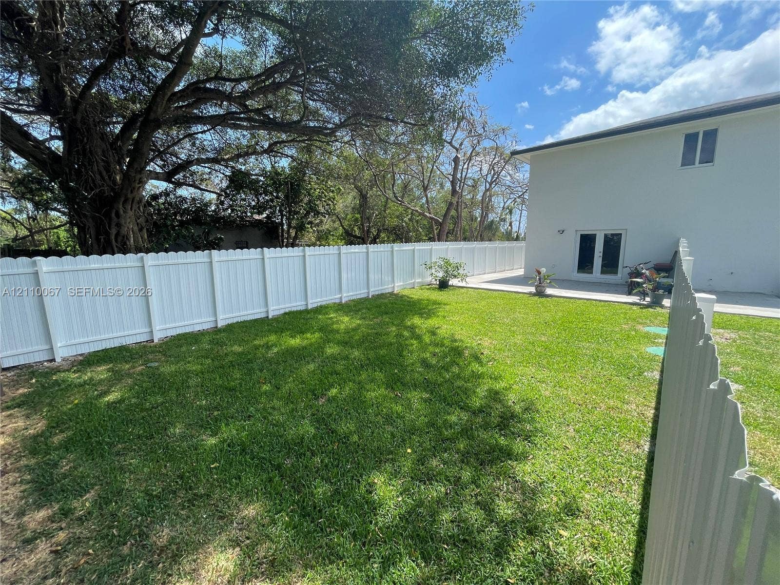 Fenced backyard featuring a manicured lawn, concrete patio, white vinyl perimeter fencing, and mature canopy trees
