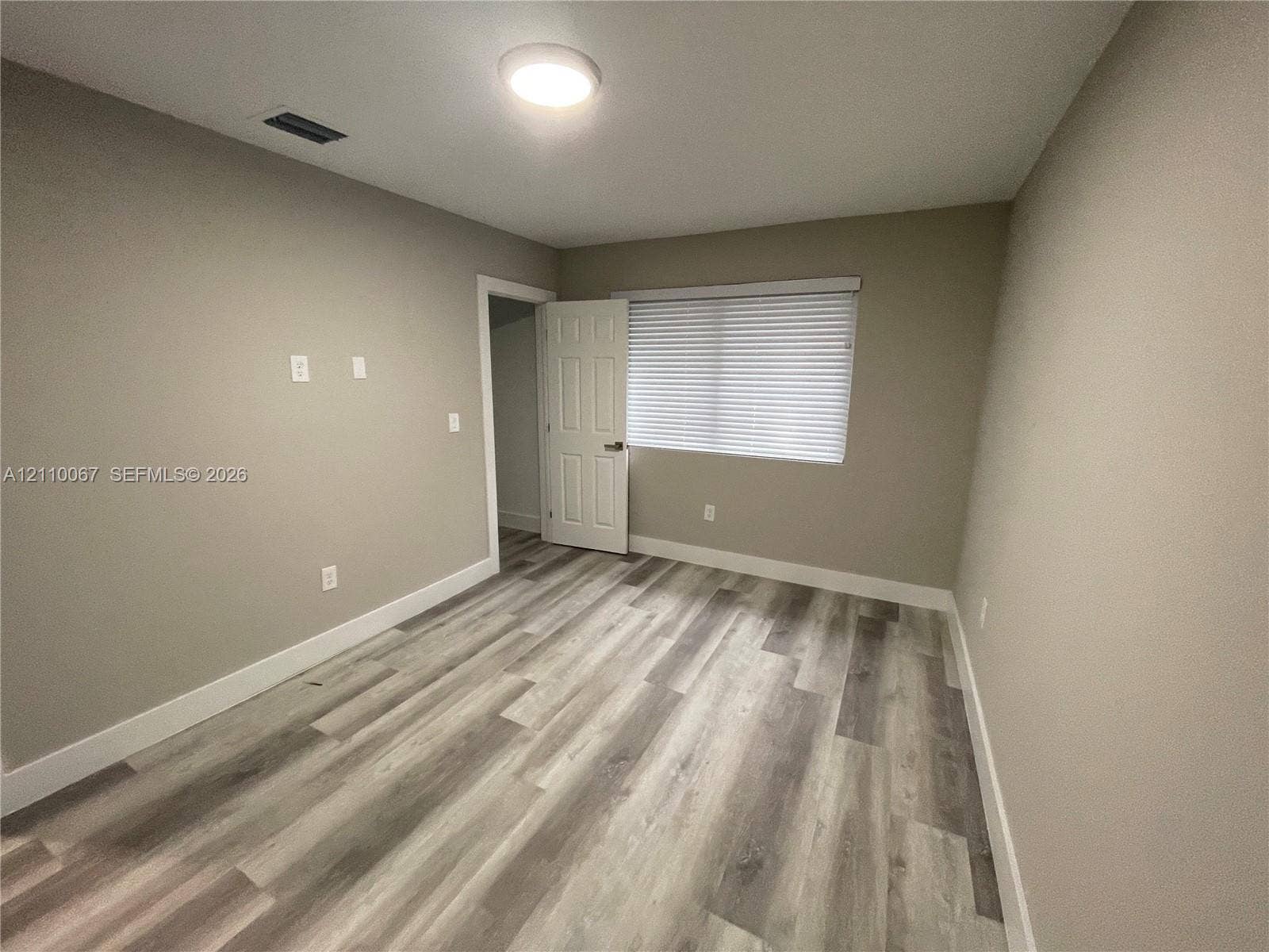 Neutral-toned interior space featuring wood-finish flooring, a six-panel door, and a horizontal window with adjustable blinds