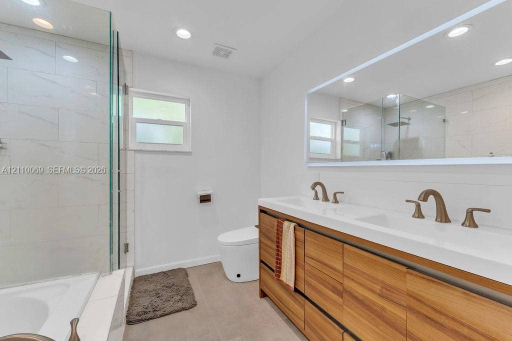 4325 NW 95th Terrace, Sunrise, FL 33351 Dual-sink vanity with wood-finish cabinetry, white countertop, brass-tone hardware, large wall-mounted mirror, and glass-enclosed shower with light-toned wall tile