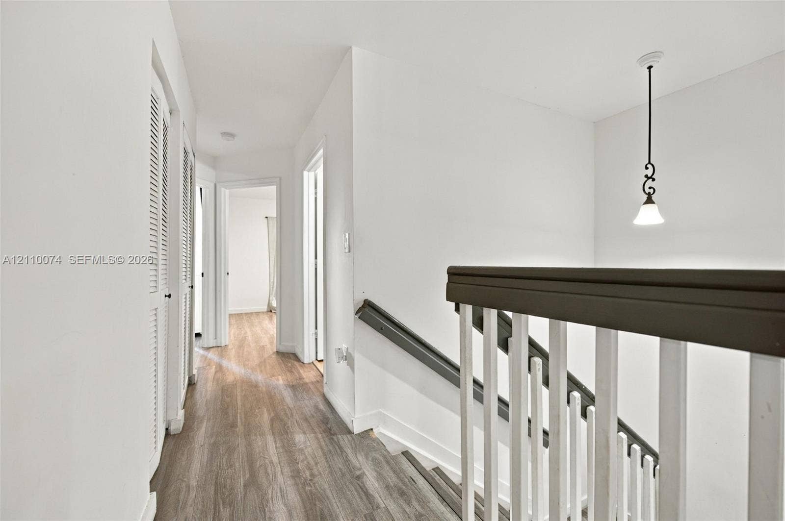 17370 NW 69th Court, #304, Hialeah, FL 33015 Hallway featuring wood-finish flooring, white louvered closet doors, and a staircase with a dark-finish handrail and white balusters, accented by a decorative hanging pendant light