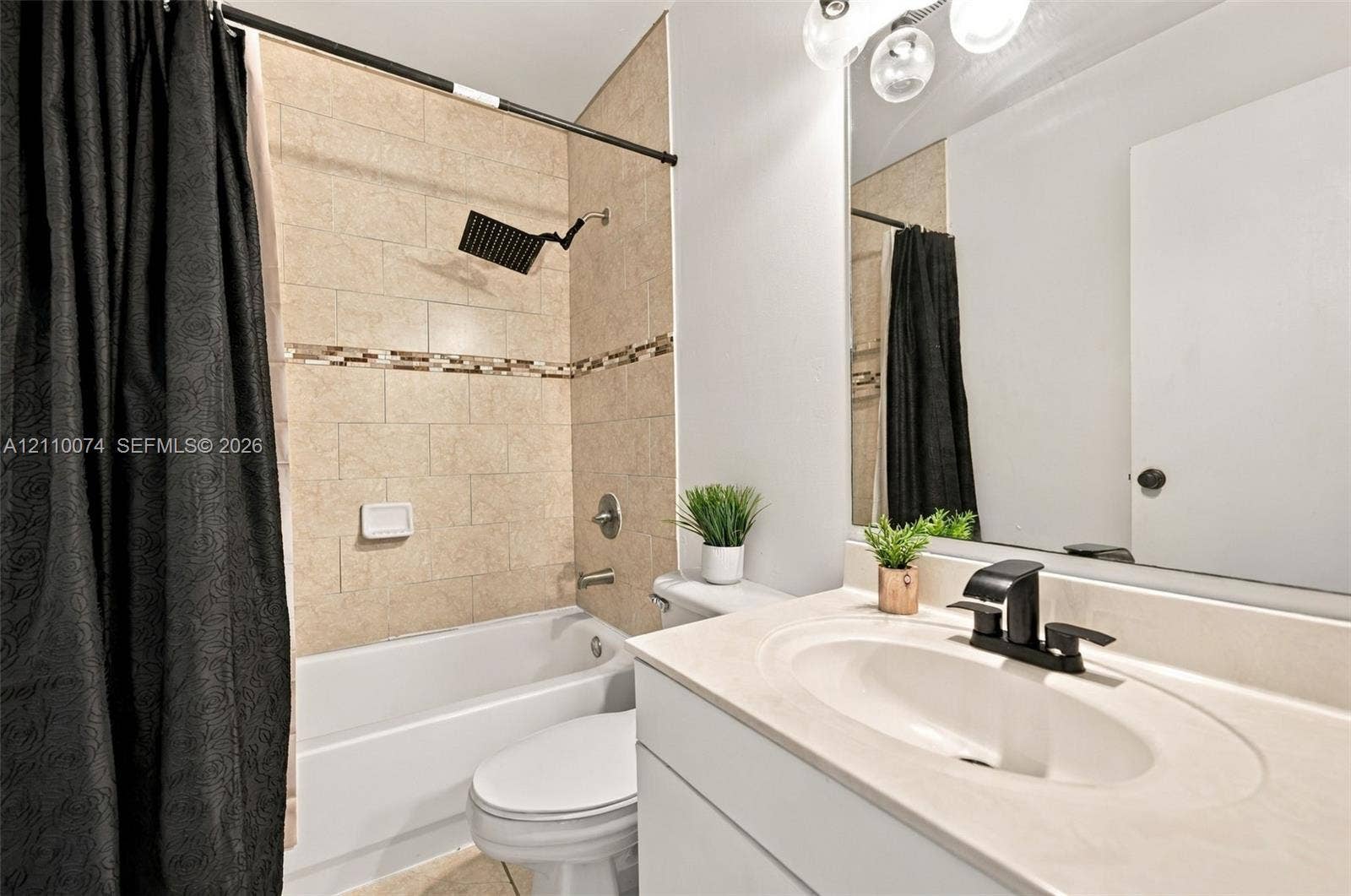 17370 NW 69th Court, #304, Hialeah, FL 33015 Full bathroom featuring a white vanity with a neutral solid-surface countertop, an integrated oval sink with a matte black faucet, a large wall-mounted mirror, and a bathtub surround tiled in neutral stone-finish ceramic with a decorative glass-tile inlay and a matte black rainfall showerhead