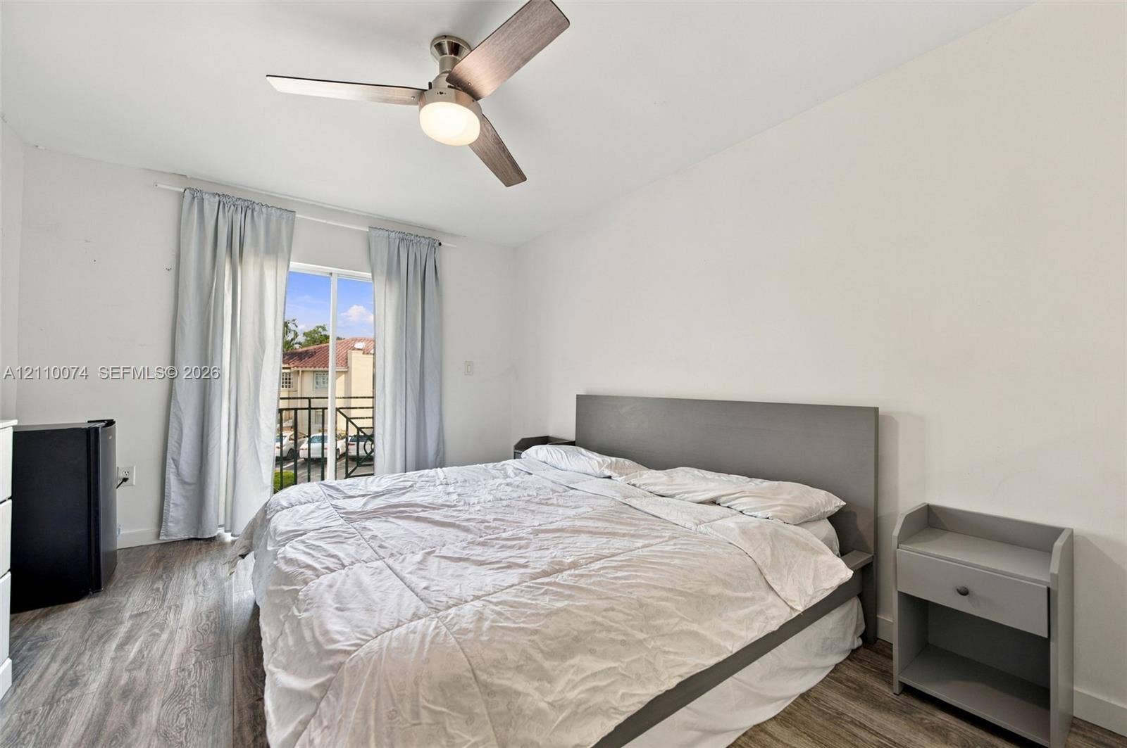 17370 NW 69th Court, #304, Hialeah, FL 33015 Neutral-toned room featuring wood-finish flooring, a contemporary three-blade ceiling fan with integrated lighting, and a sliding glass door providing external access to a balcony with metal railings