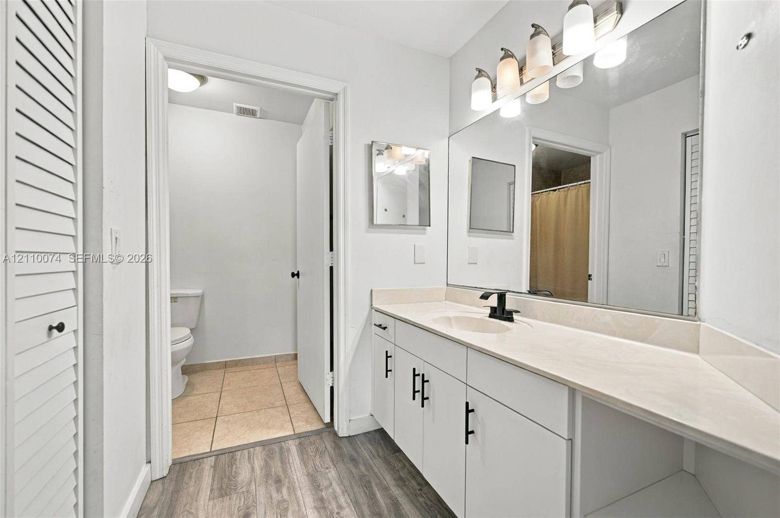 17370 NW 69th Court, #304, Hialeah, FL 33015 Bathroom vanity featuring a light-toned solid surface countertop, matte black faucet, and white shaker-style cabinetry with matte black hardware, paired with a full-width vanity mirror and wood-finish flooring