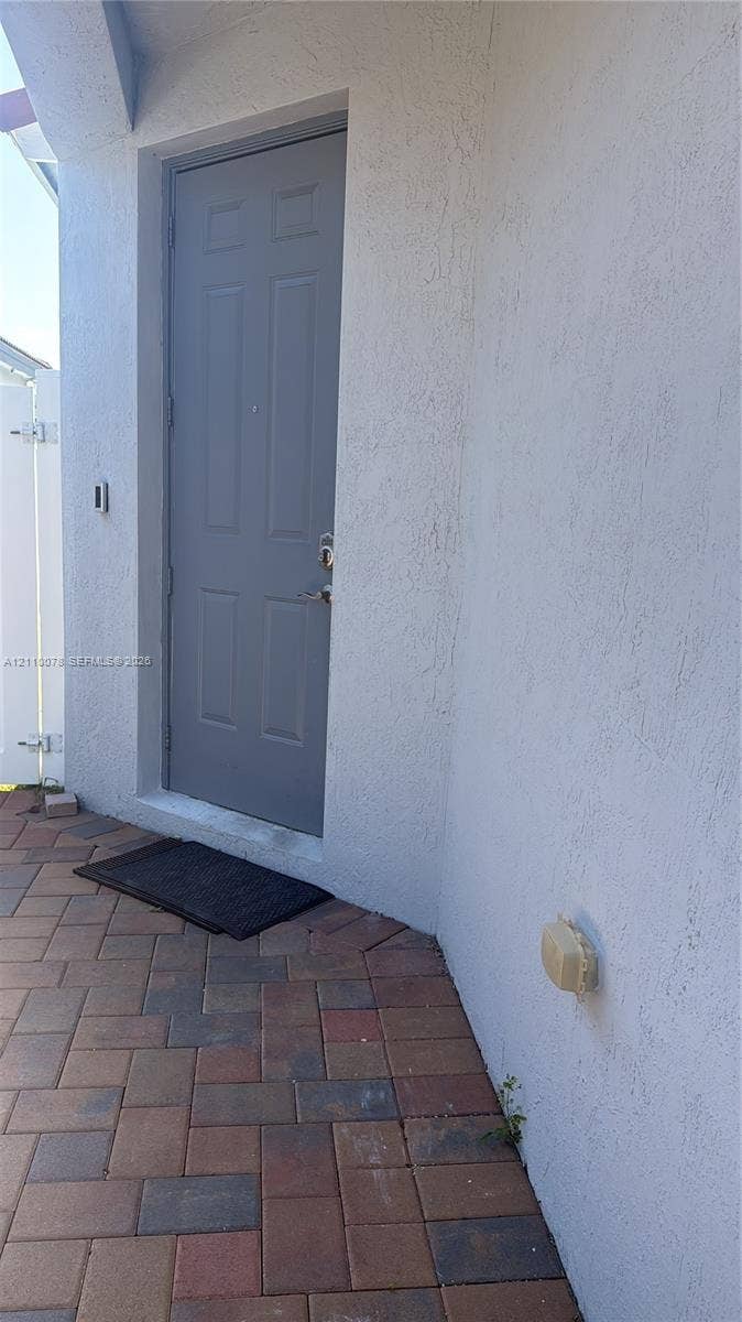 Exterior entryway featuring a textured stucco finish, multi-panel gray door, and brick-style paver flooring
