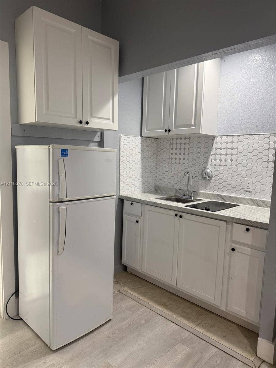 White cabinetry with matte black hardware, stone-finish countertops, hexagonal tile backsplash, stainless steel sink, and integrated stovetop
