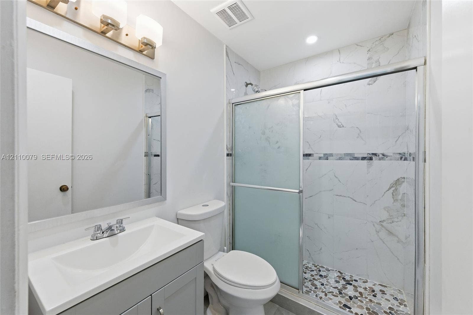 4106 NW 88th Avenue, #3, Coral Springs, FL 33065 Bathroom featuring a gray vanity with a white countertop, framed wall mirror, three-light fixture, and a glass-enclosed shower with tiled walls and pebble-stone flooring