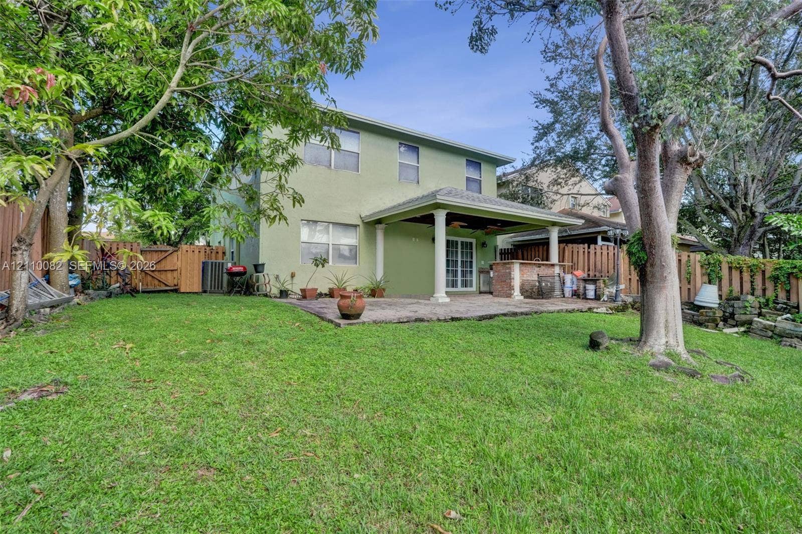Rear exterior featuring a covered patio, built-in masonry outdoor kitchen, grass lawn, wood privacy fencing, and mature trees