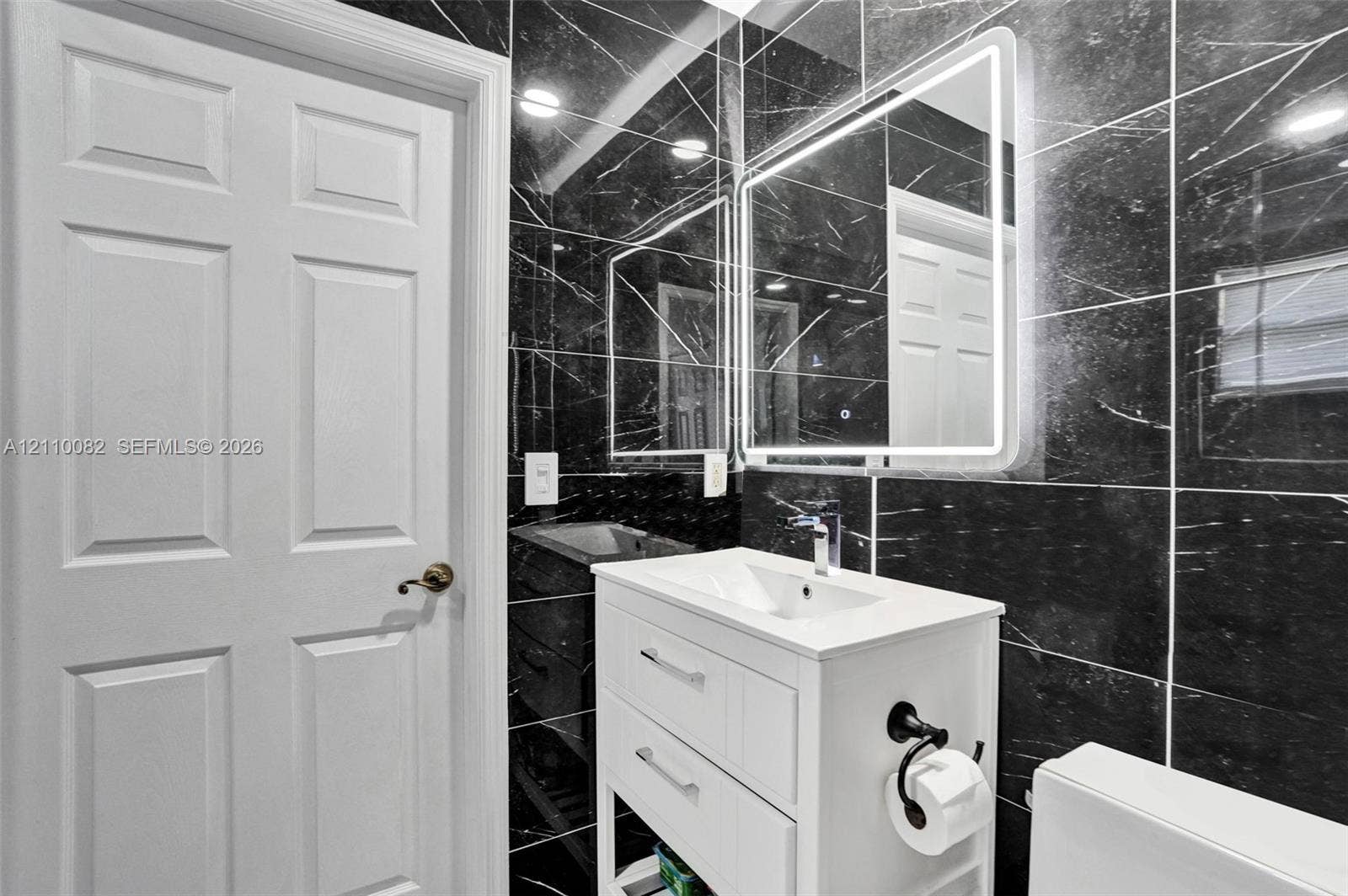 White vanity cabinet with an integrated basin, sleek chrome faucet, LED-backlit mirror, and full-height black stone-finish wall tile