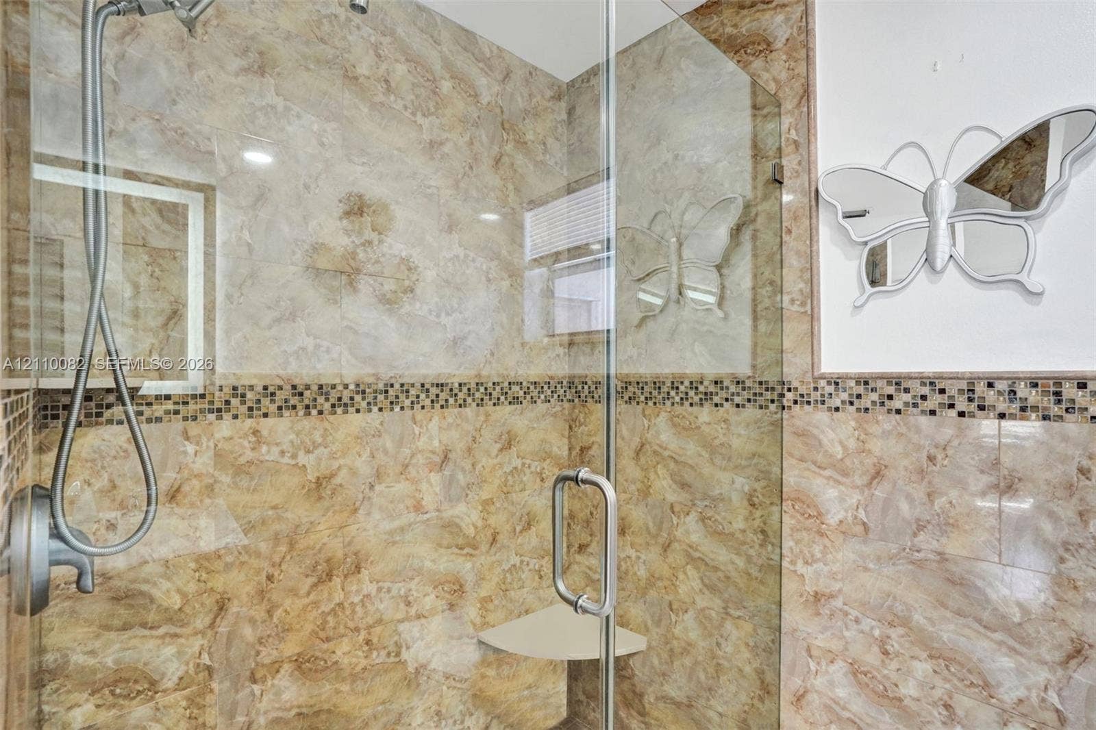 Walk-in shower featuring floor-to-ceiling stone-finish tile, glass enclosure, handheld shower fixture, built-in corner shelving, and decorative mosaic accent tile inlay