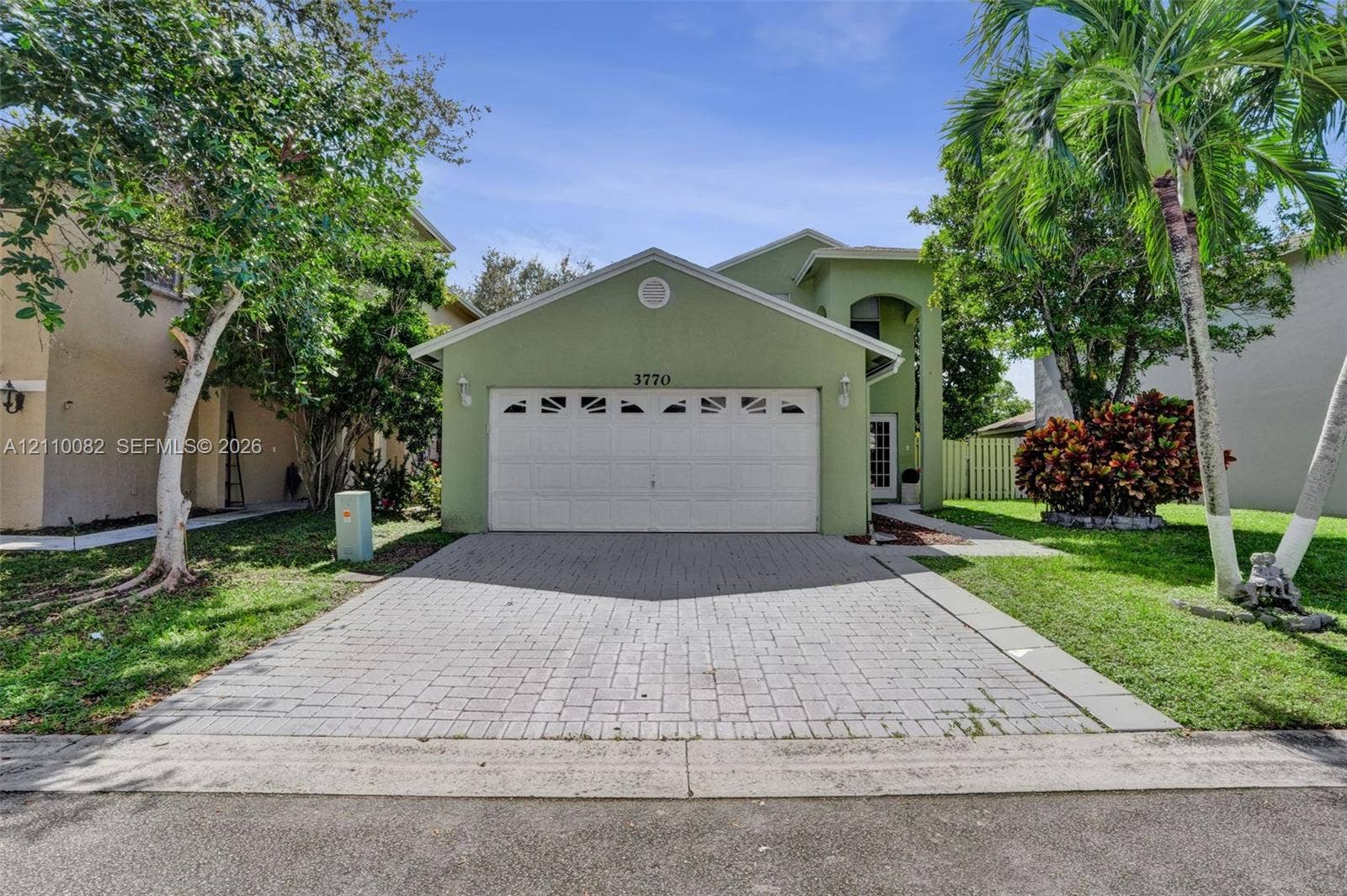 Stucco exterior featuring a dual-car garage with arched decorative glass inserts and a paved stone driveway, surrounded by mature palm trees and landscaped shrubbery
