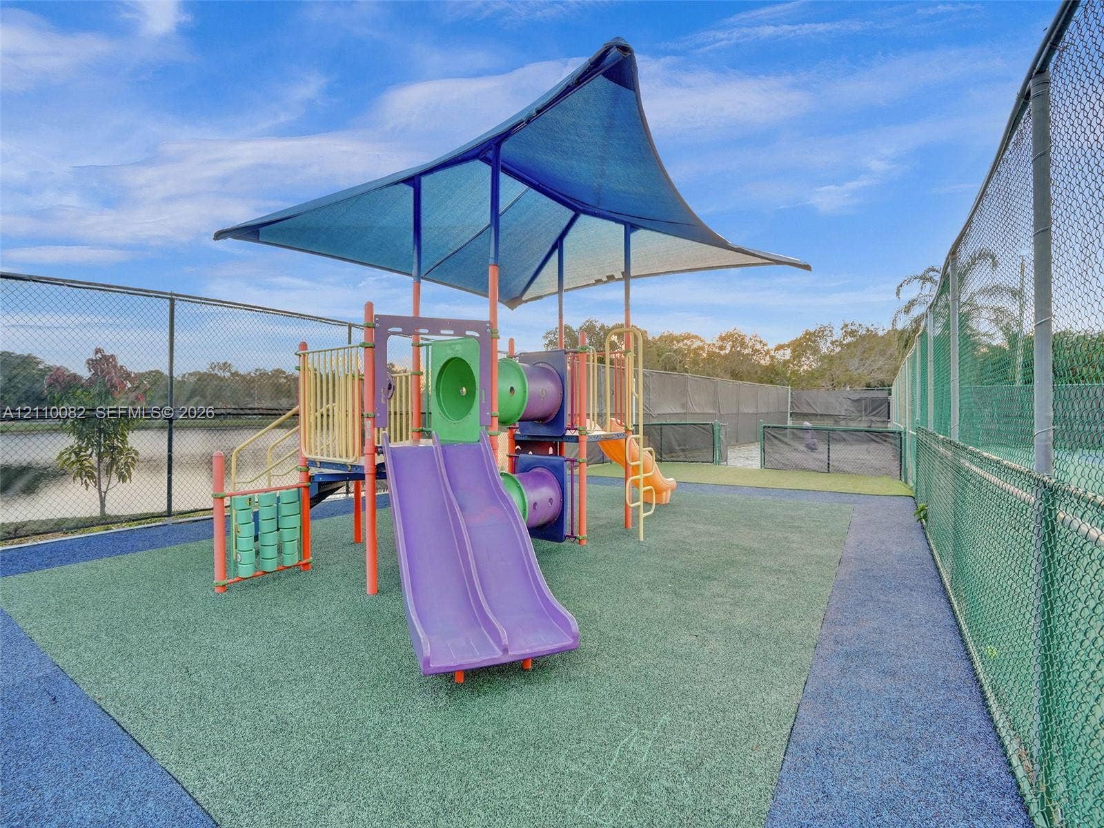 Recreational area featuring a playground structure with dual slides, a protective shade canopy, impact-absorbing surfacing, and perimeter chain-link fencing